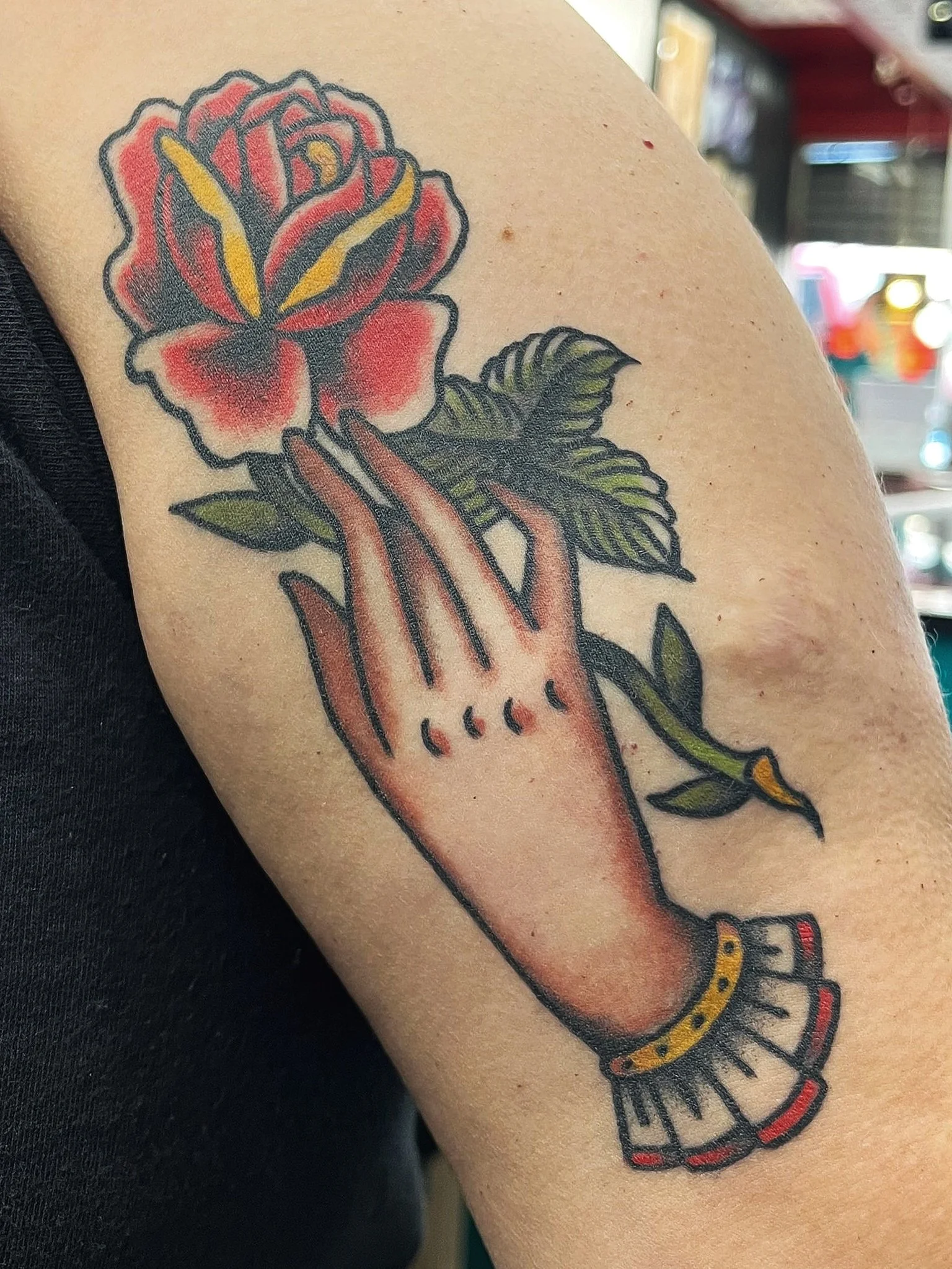 Tattoo of a hand with a ruffled cuff reaching towards a red and pink flower with green leaves.