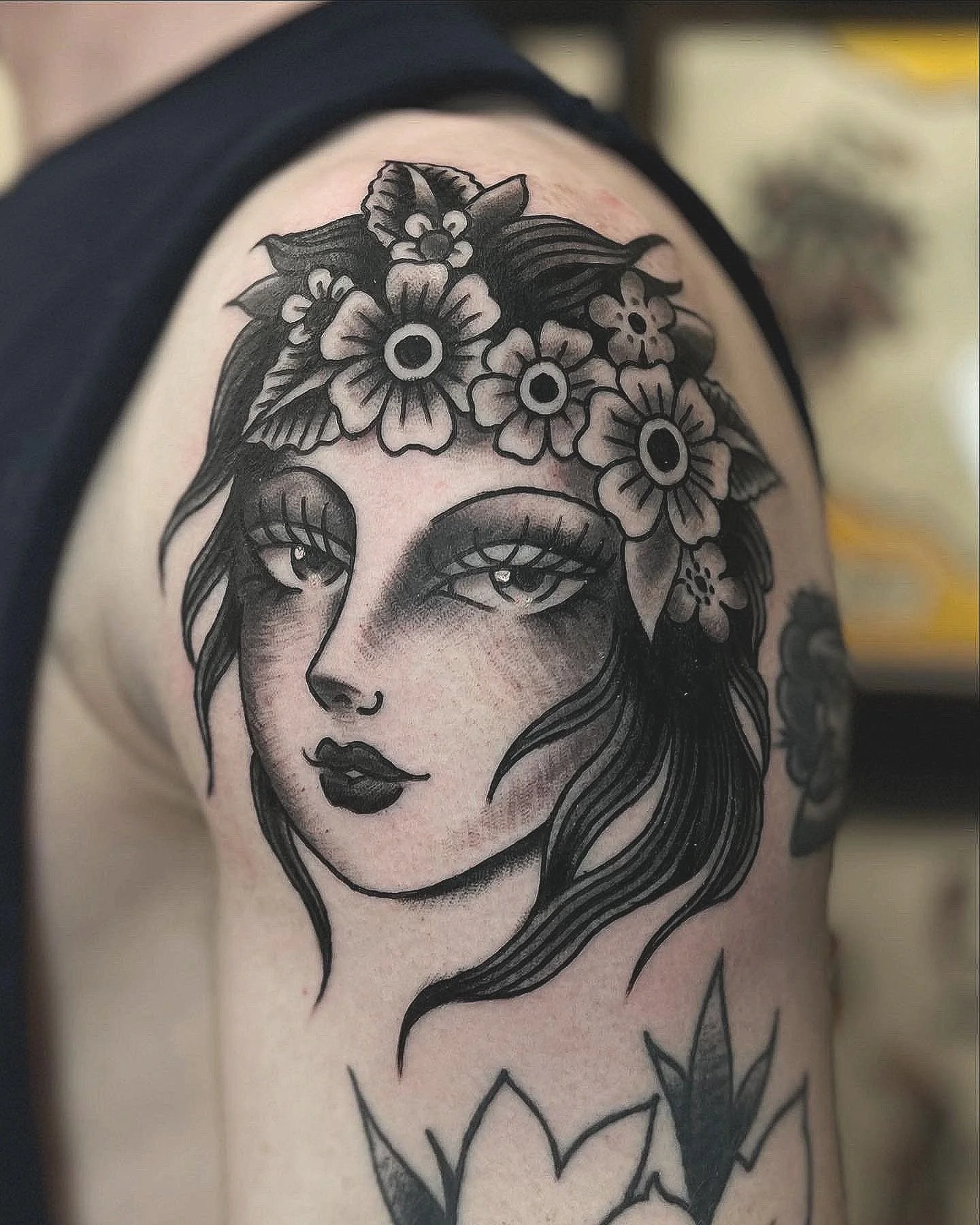 Black and gray tattoo of a woman's face with flowing hair and a floral crown on her head, featuring large eyes and dark lipstick.