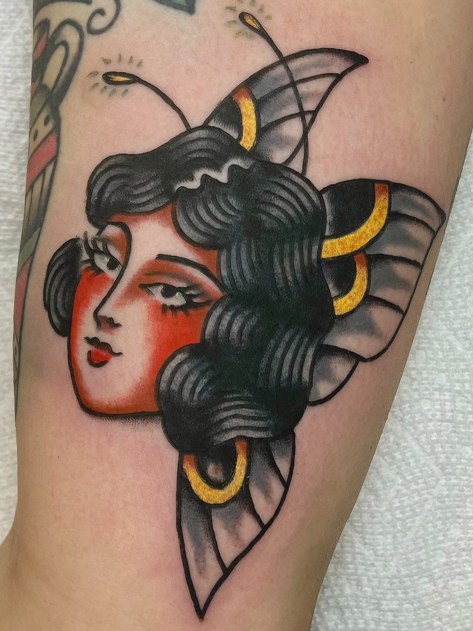 Tattoo of a woman with dark hair and red cheeks, with a butterfly wing behind her head, decorated with gold accents, on a person's arm.
