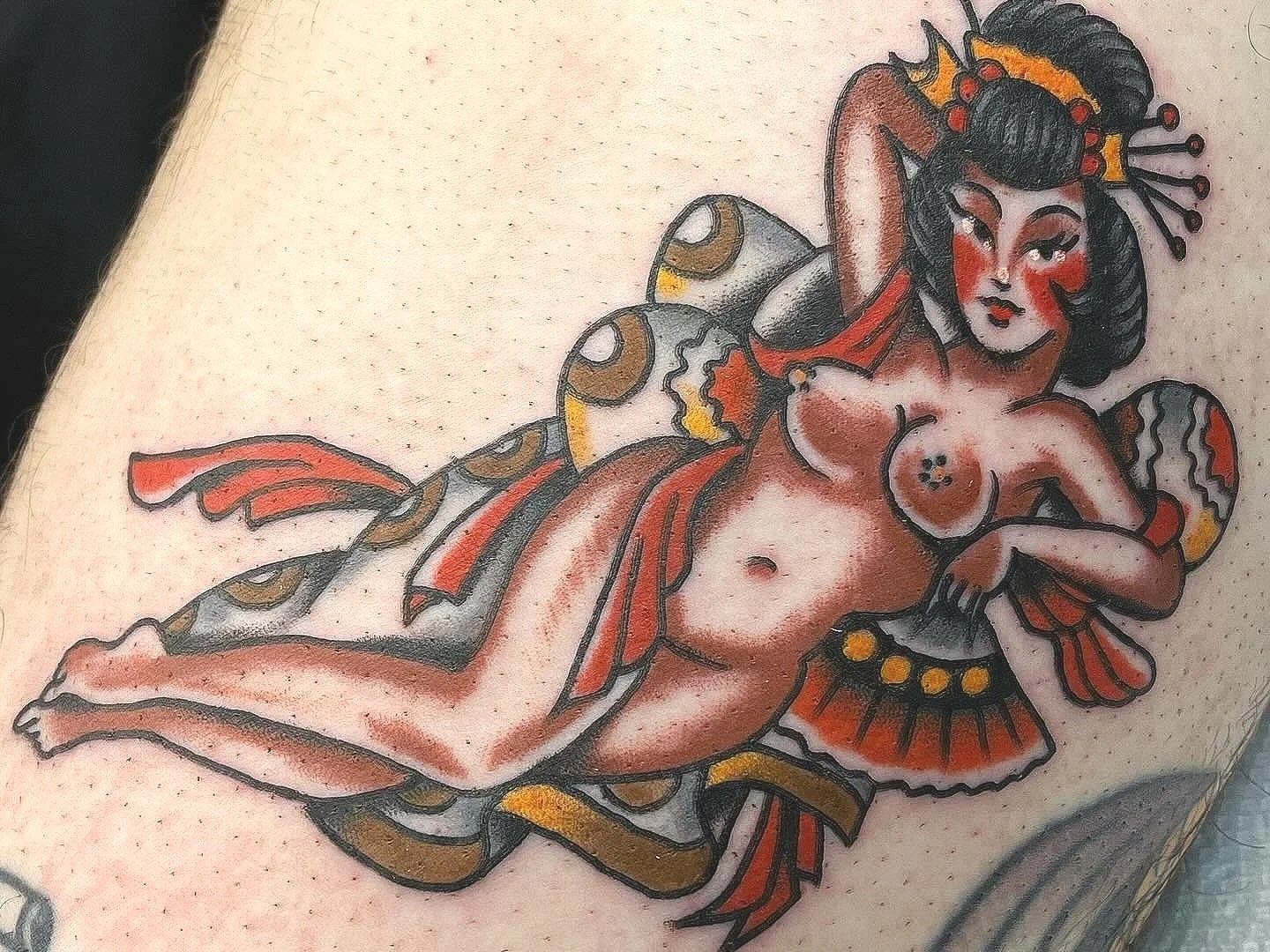 Tattoo of a stylized woman with black hair and a crown, reclining on a fish with orange, black, and gray scales, holding a fan, in traditional Japanese tattoo style.