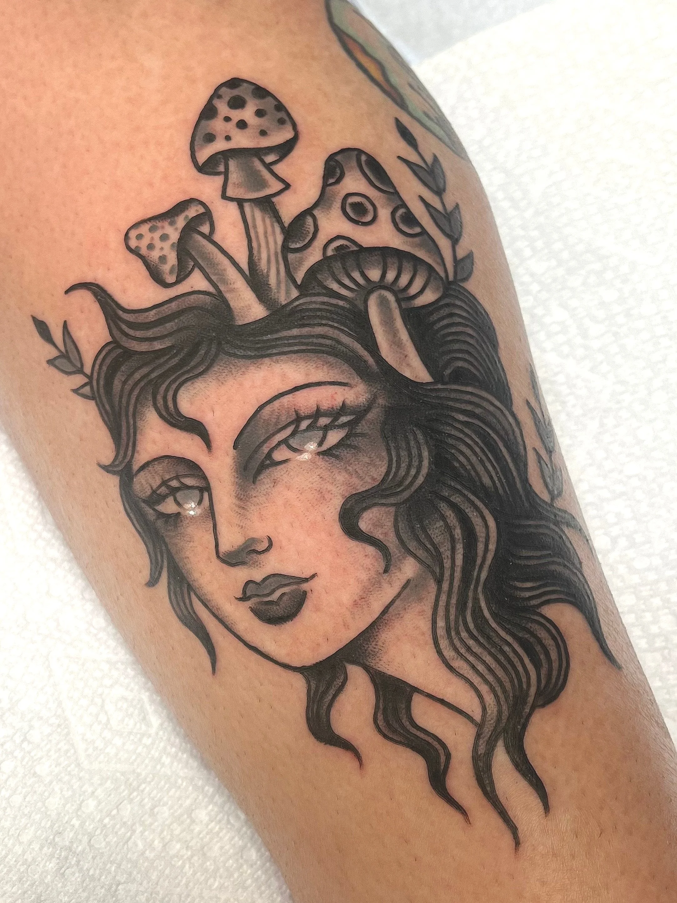 Tattoo of a woman's face with flowing hair, surrounded by mushrooms and leaves.