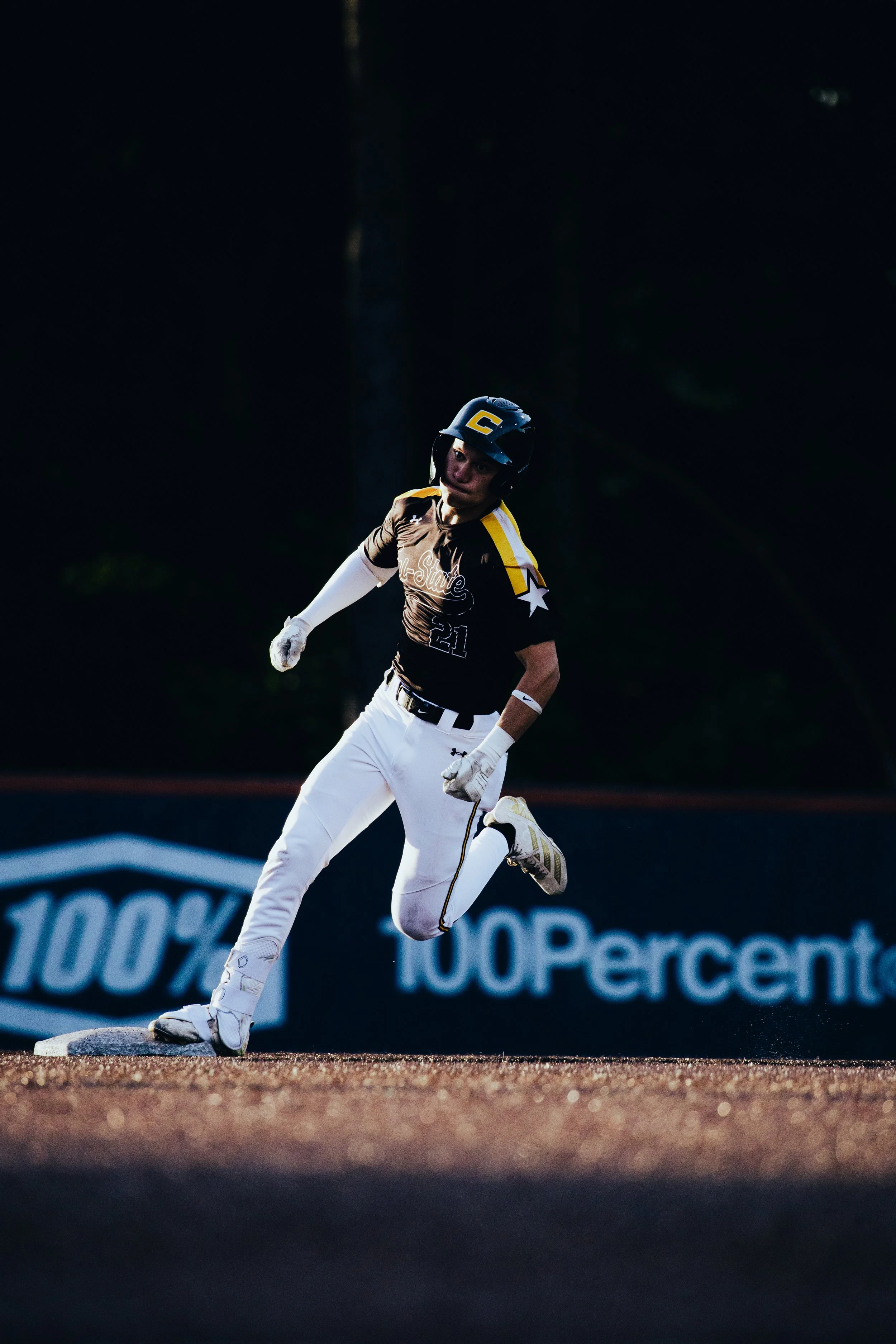 A baseball player in a brown jersey and white pants is running on the field, wearing a black helmet. The background is dark with advertising banners.