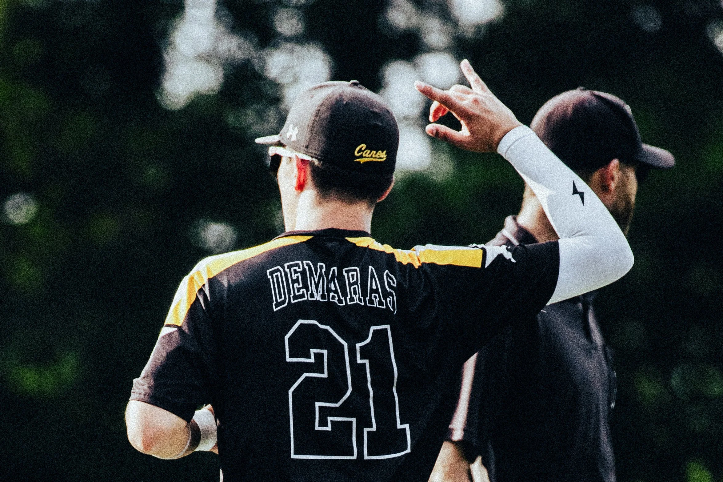 A baseball player wearing a cap with the word 'Canea' and a jersey with the name 'DeMaras' and number 22, is making a gesture towards another person during outdoor evening or late afternoon.