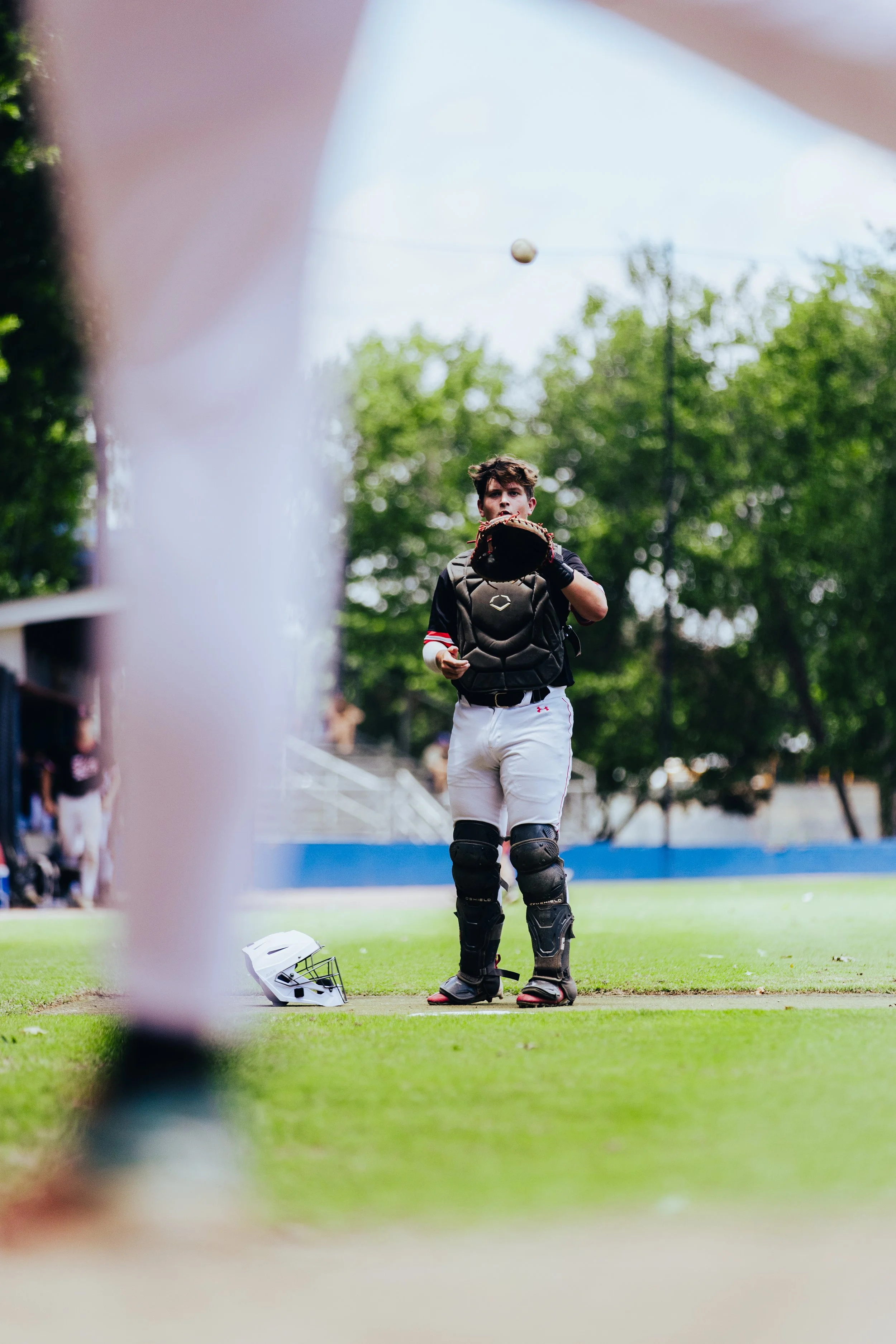 A young baseball catcher standing on the field in uniform with a helmet on the ground in front of him, preparing to catch a baseball.