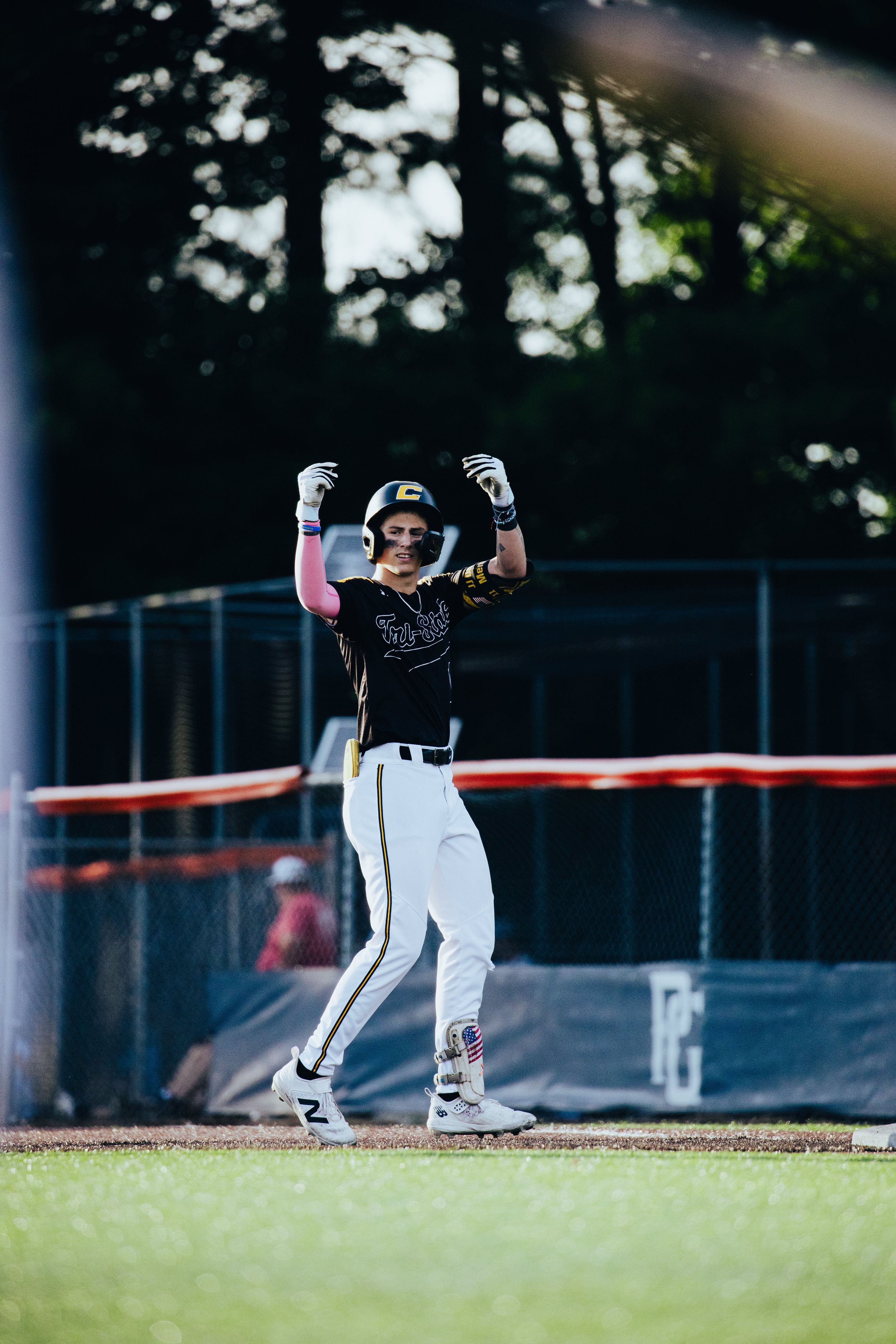 A baseball player in a black jersey and white pants standing on the field, wearing a helmet, gloves, and cleats, with his arms raised in celebration.