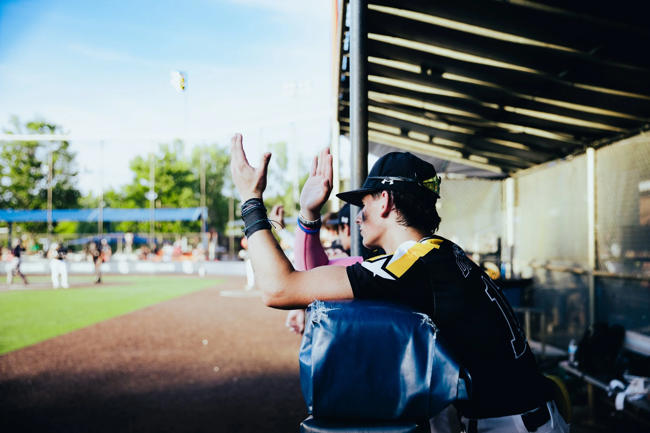 A young baseball player wearing a cap and jersey sits on the bench giving a high-five to a teammate during a game, with a baseball field and spectators in the background under a clear blue sky.