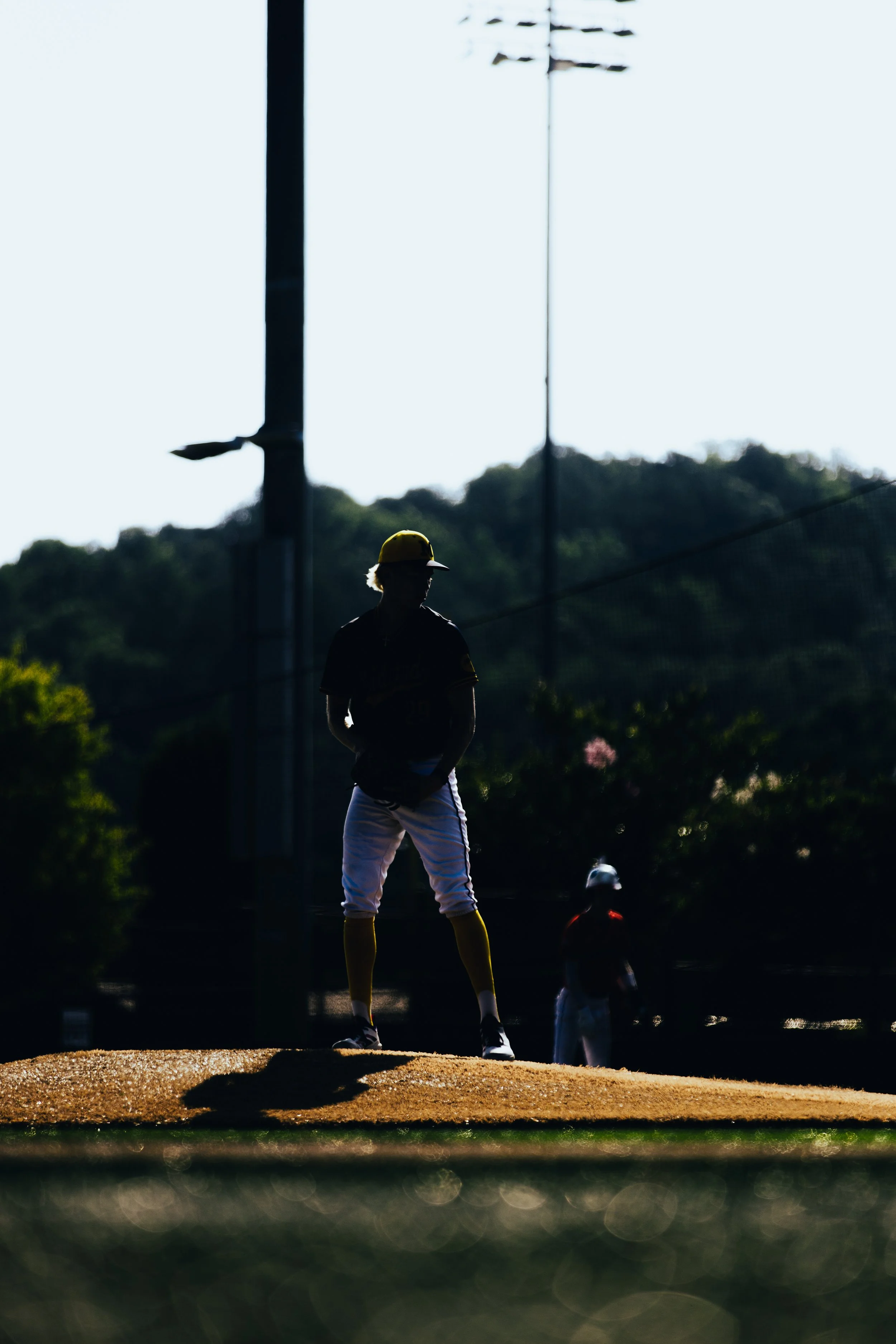 Silhouette of a baseball player in uniform and yellow cap standing on a baseball field, with other players and a faint background of power lines and trees.