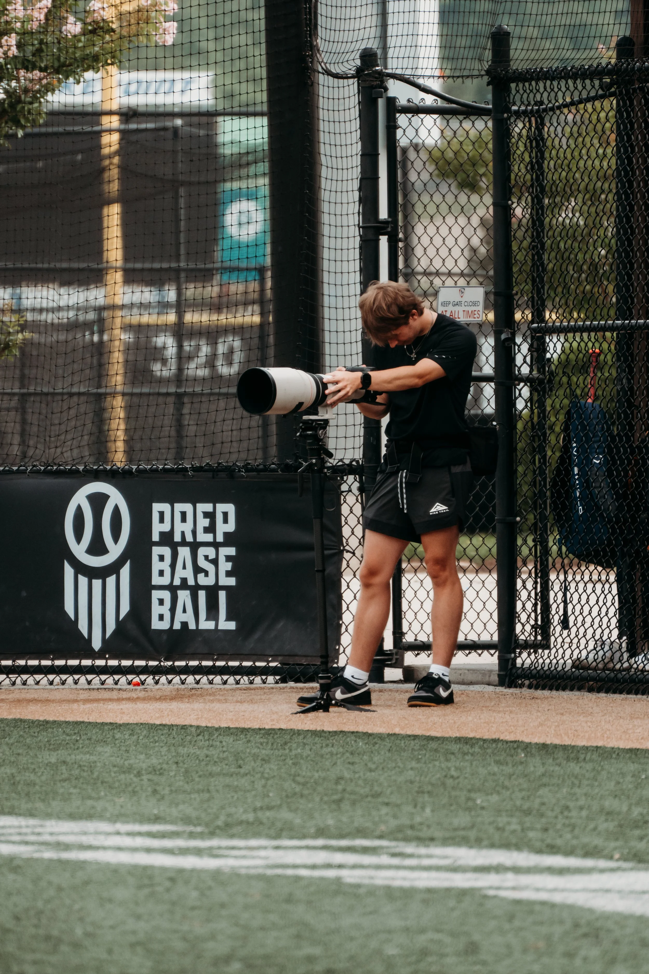 A person with short brown hair, wearing a black t-shirt, black shorts, and Nike sneakers, adjusts a large camera lens mounted on a tripod near a chain-link fence at a baseball field.