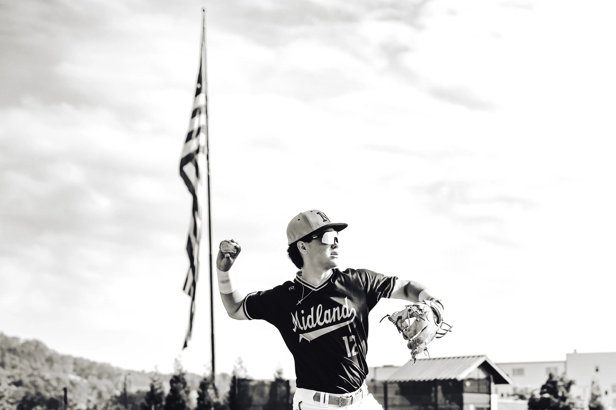 A young man wearing sunglasses, a baseball cap, and a Midland sports jersey is playing baseball outdoors. He has his right arm raised with a clenched fist and is holding a baseball glove in his left hand. The scene is set against a cloudy sky, with s