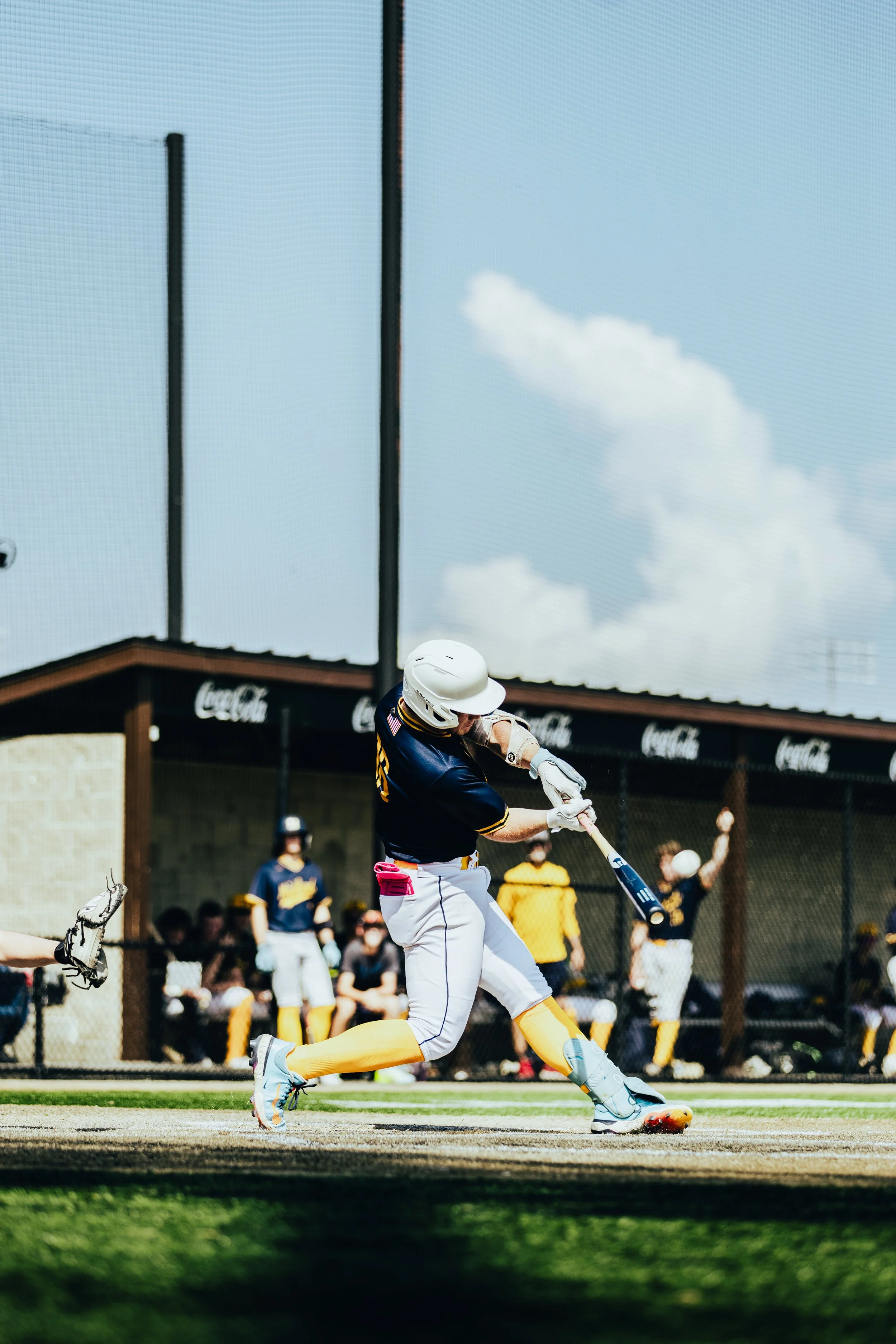 A baseball player in a navy and yellow uniform swings at a pitched ball during a game, with other players and spectators visible in the background.