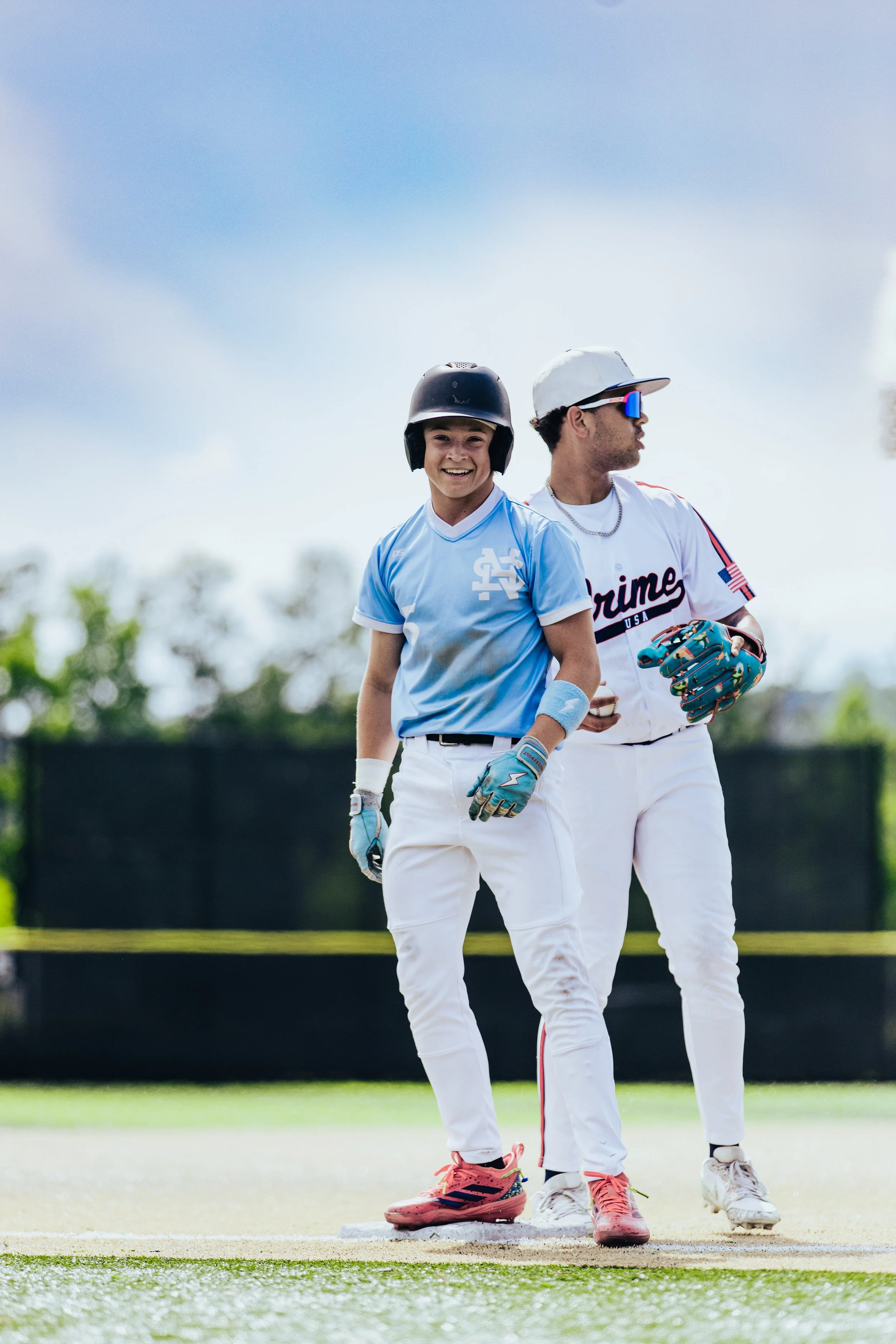 Two young boys playing baseball outdoors on a sunny day, one smiling at the camera and the other looking to the side, both wearing baseball gloves and helmets.