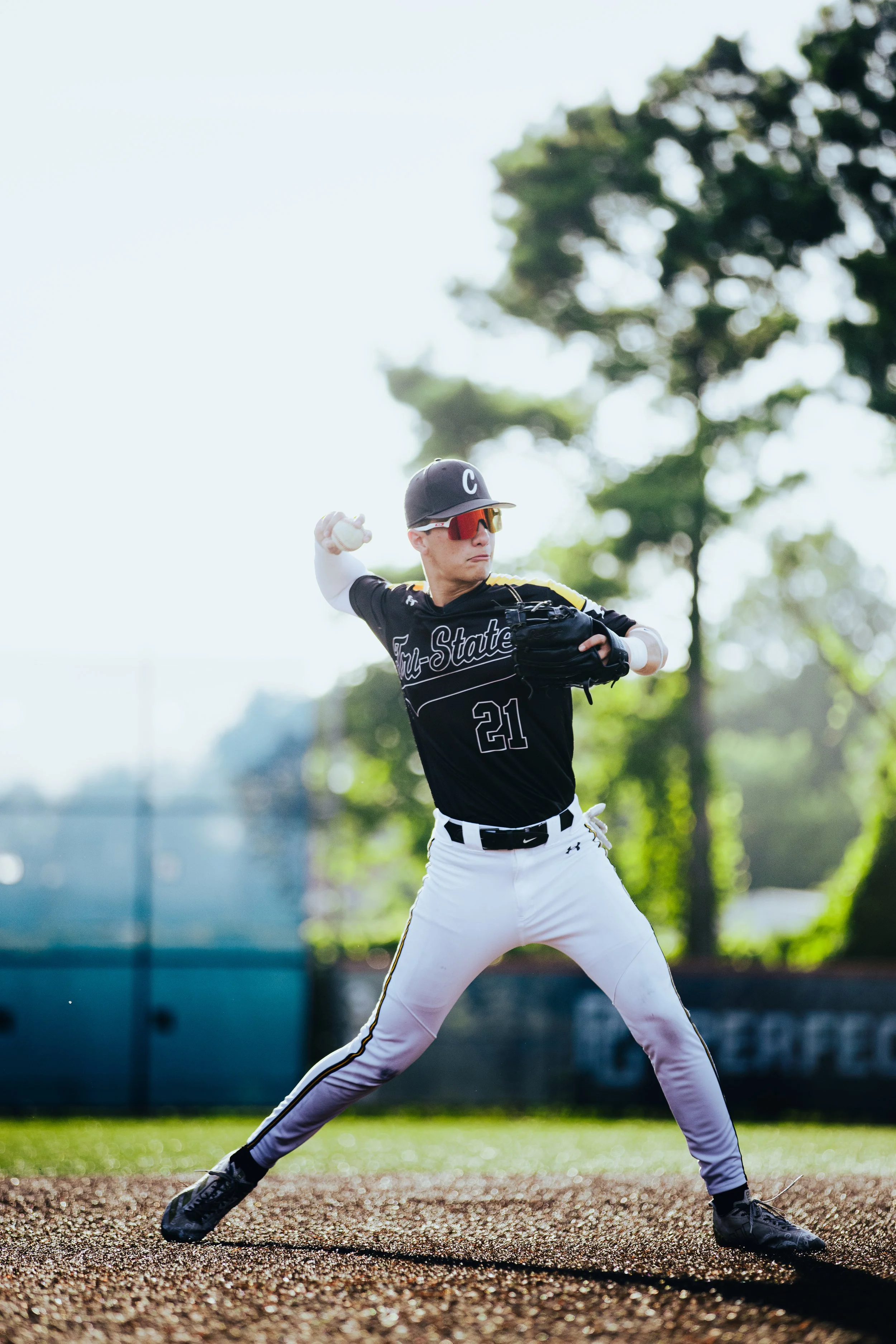 Baseball player in black jersey and white pants in a pitching stance, outdoors on a baseball field with trees in the background.