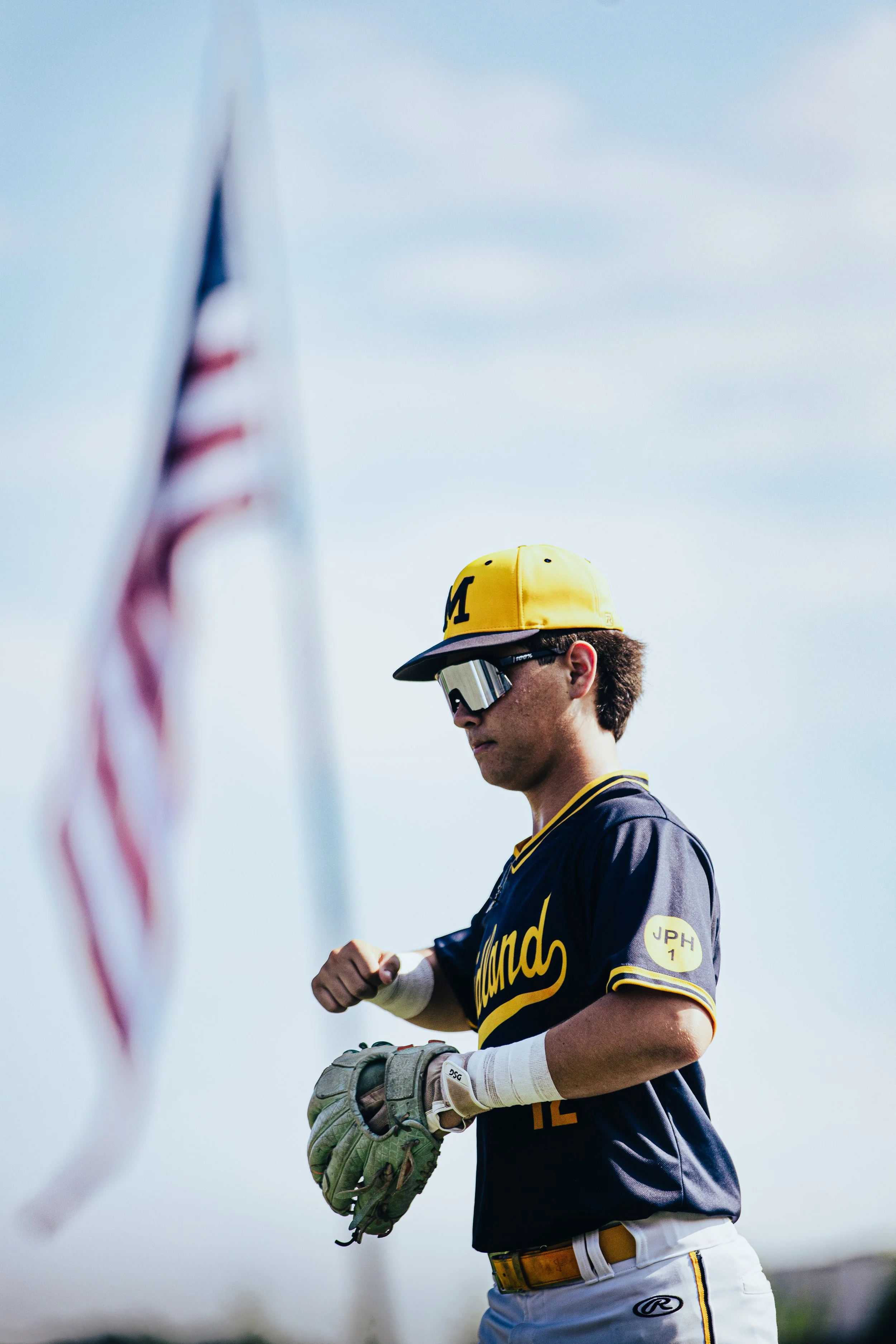 A baseball player wearing a navy blue and yellow uniform, a yellow cap, sunglasses, and holding a baseball glove, is seen standing outdoors against a cloudy sky. Part of an American flag is visible in the foreground on the left side.