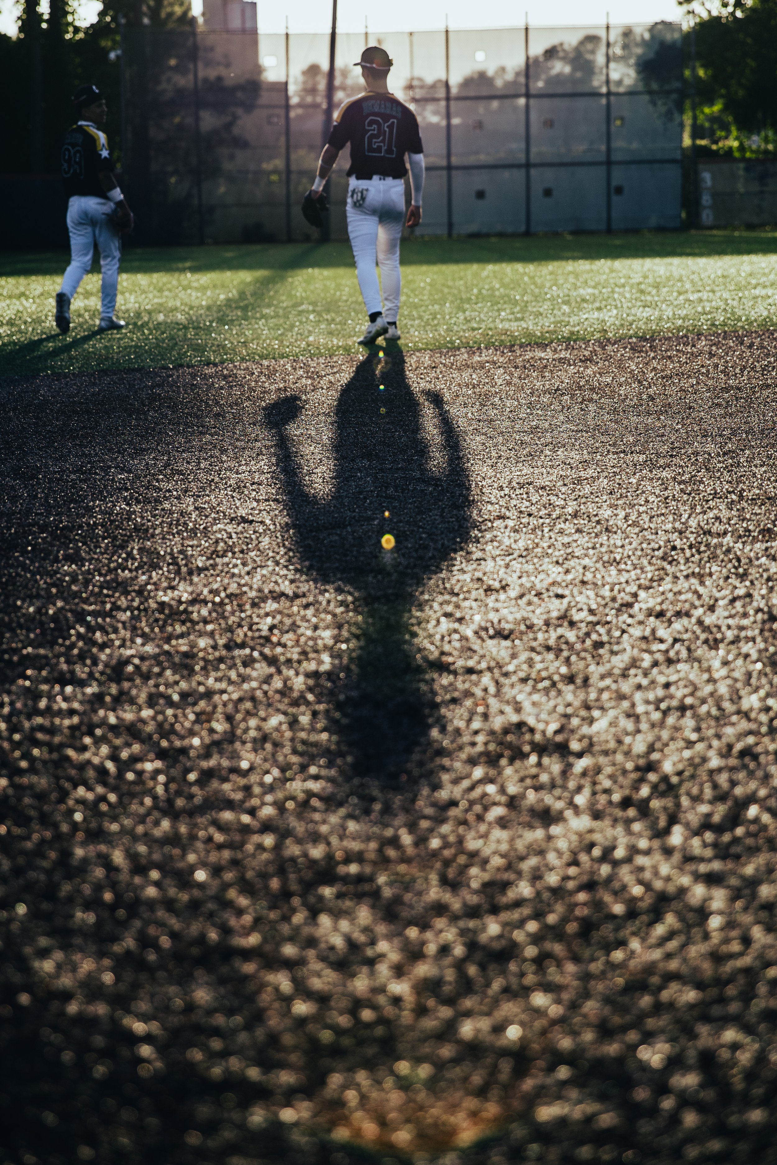 Two baseball players standing on the field with the sun setting behind them. The player in the foreground faces away, while the other is turned slightly to the side. Shadows are long, and the field has a shiny, textured surface.
