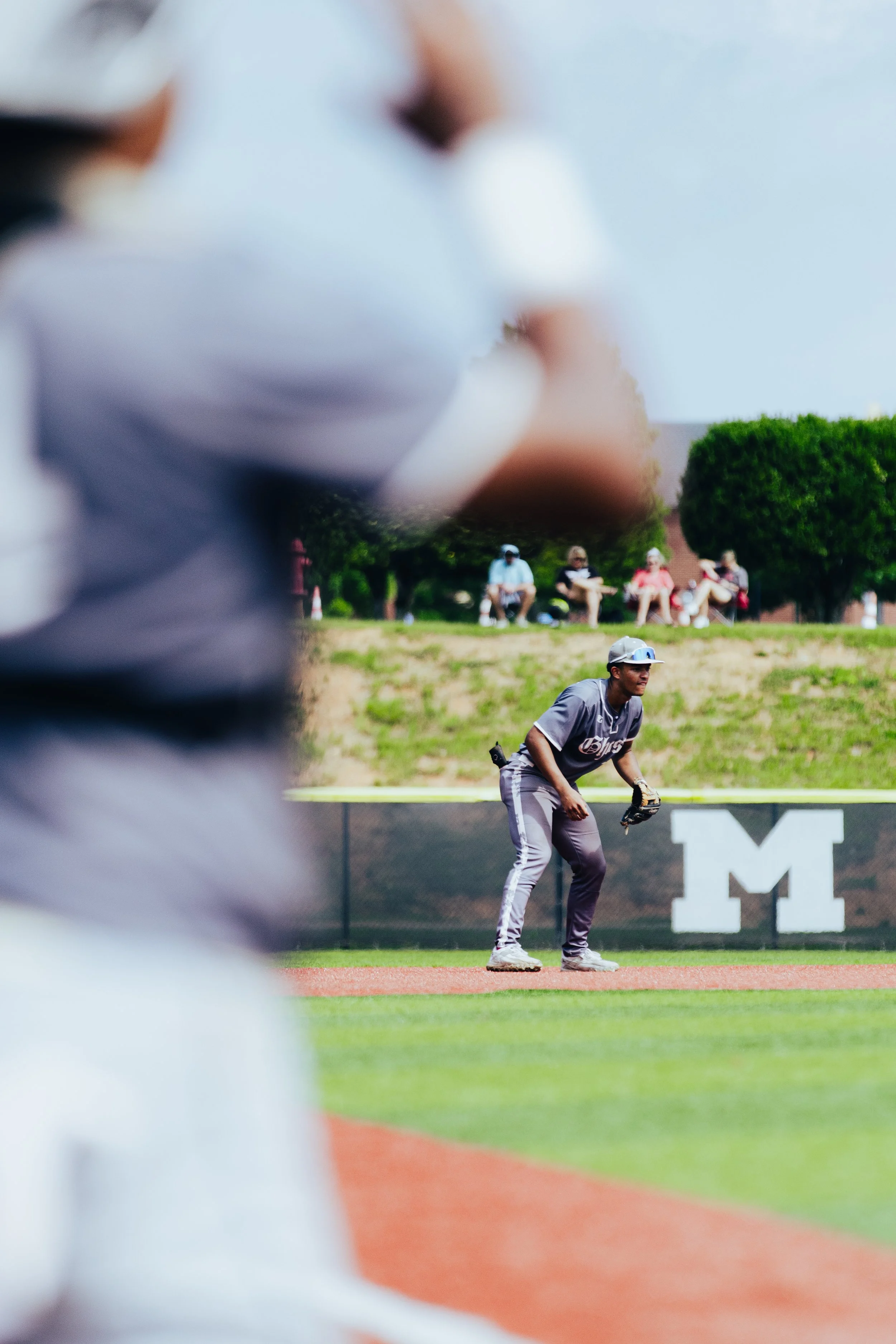 A baseball player in gray uniform and cap getting ready to field a ball on a baseball field, with spectators sitting on a grassy hill in the background.