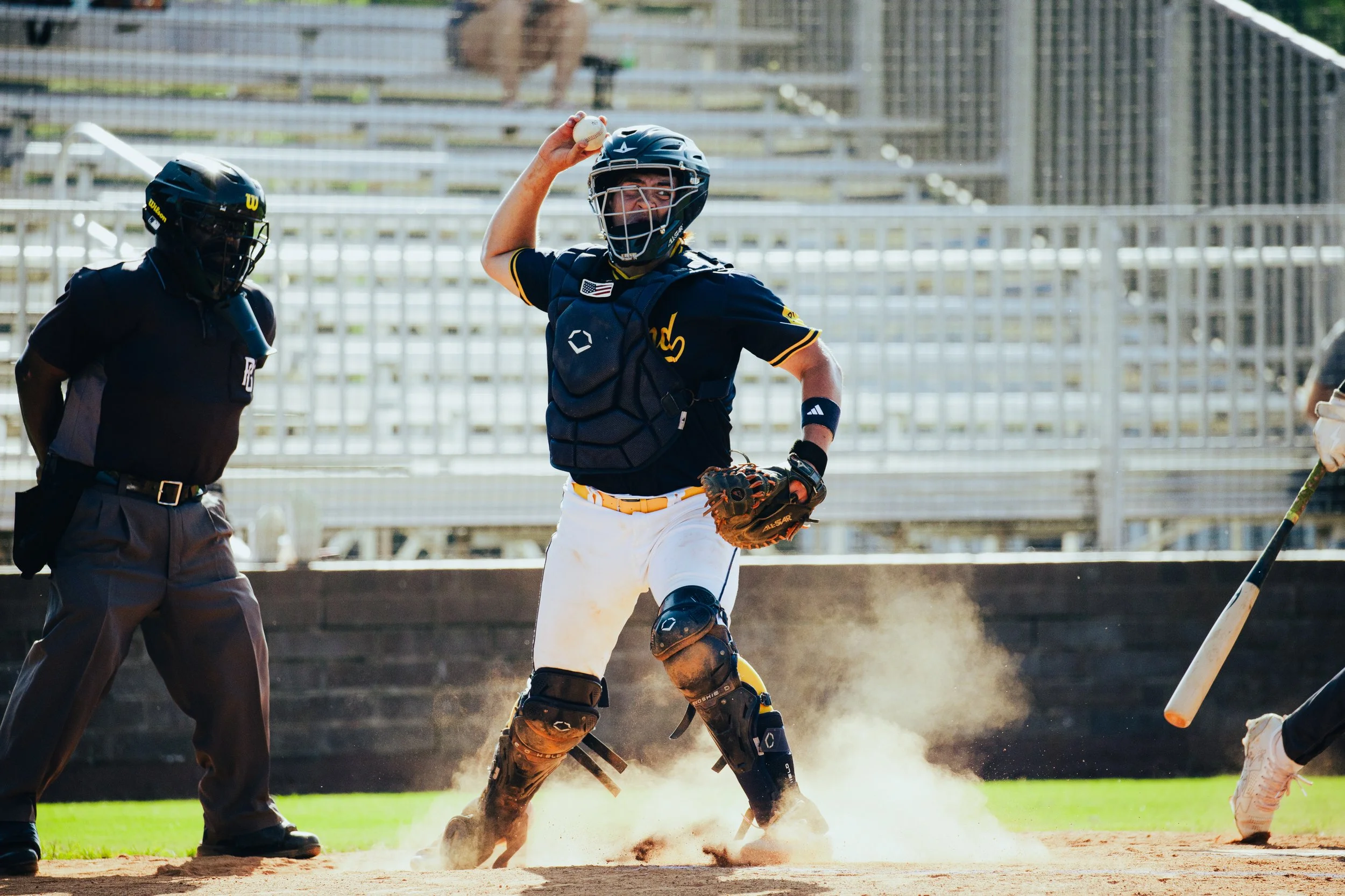 A baseball player in catcher's gear holding a baseball in the air during a game, with an umpire nearby and a dusty field in the background.