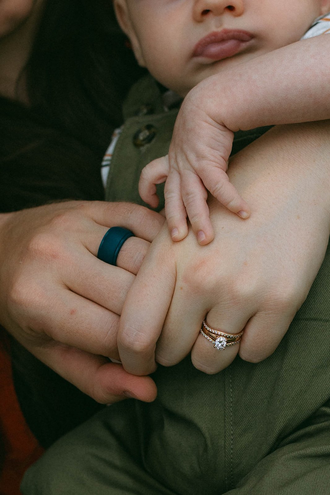 A child's hand rests on an adult's hand, which is holding the child's arm. The adult's hand has multiple rings, including a diamond ring on the ring finger. The adult's hand also has a black ring on the middle finger. The child's face is partially vi