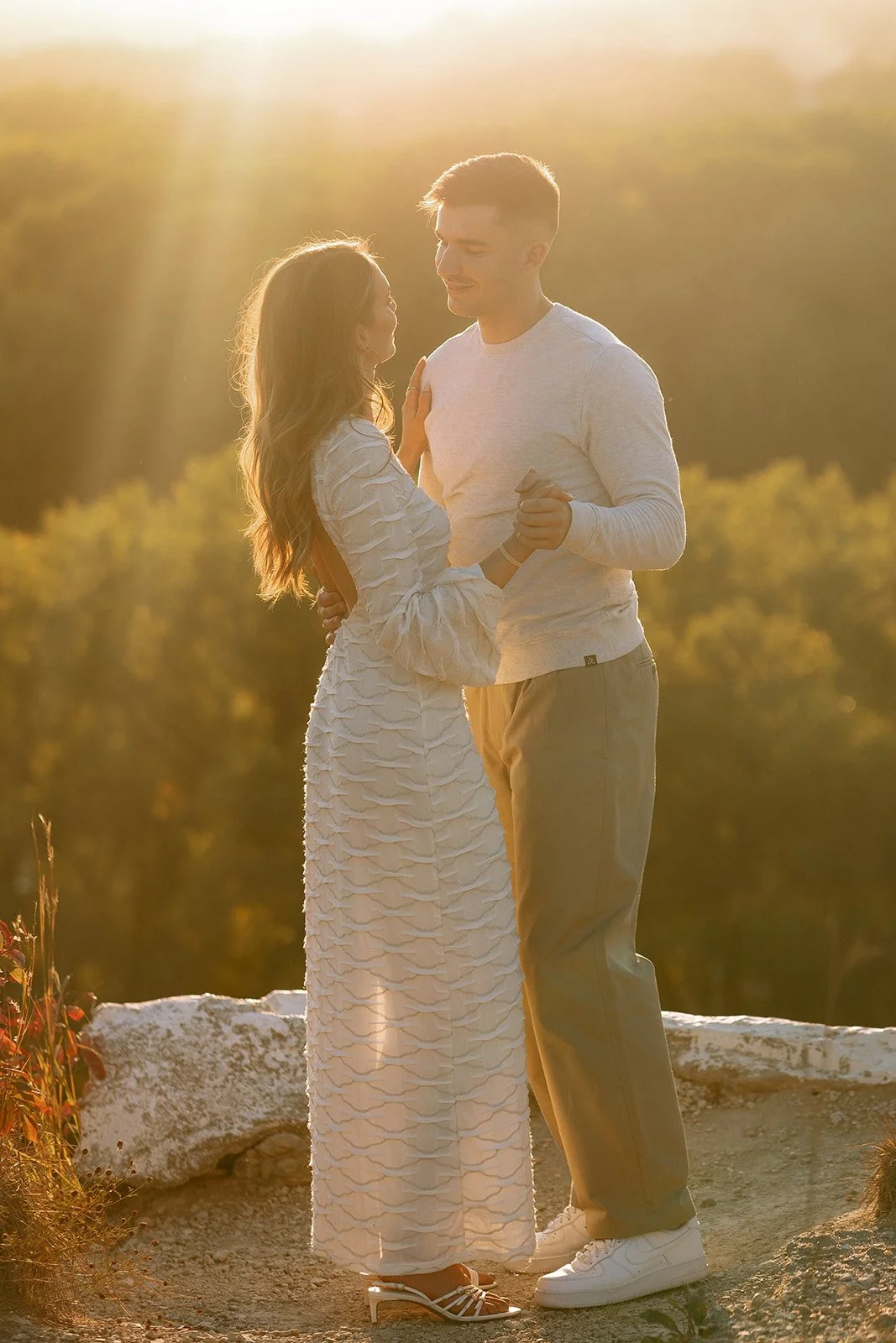 A couple stands close together outdoors at sunset, holding hands and gazing at each other, with backlit by golden sunlight.
