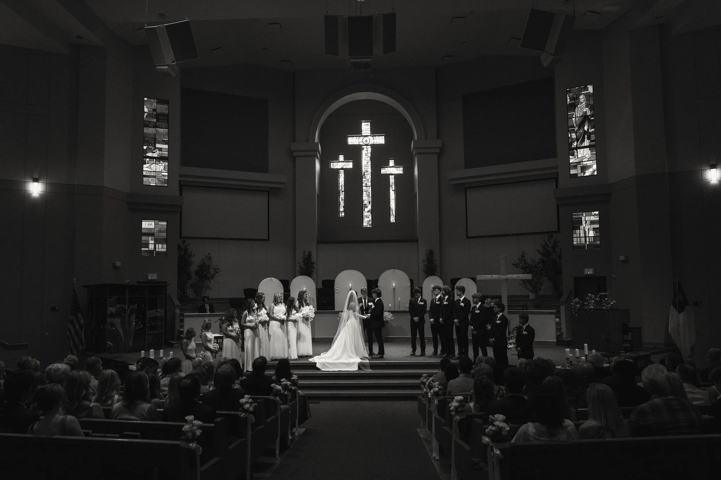 A wedding ceremony taking place inside a church with stained glass windows, where a bride and groom are standing in front of an altar surrounded by bridesmaids and groomsmen.