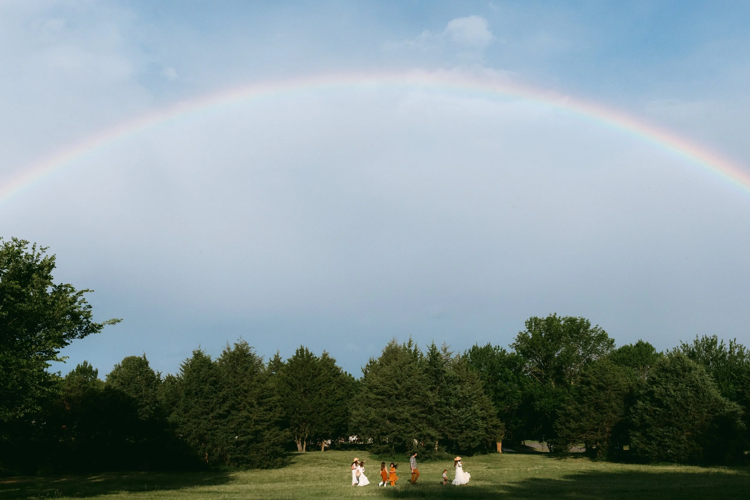 A rainbow arching across the sky over a park with tall trees and a group of people, including women in white dresses and children, gathering on the grass.