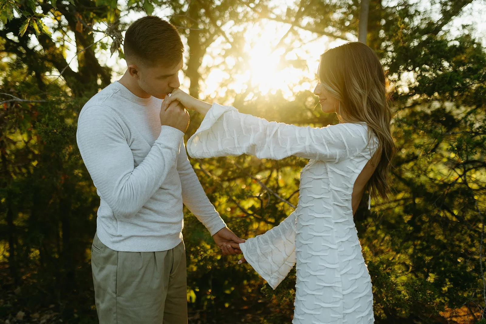 A couple holding hands outdoors at sunset, with the woman touching the man's lips.