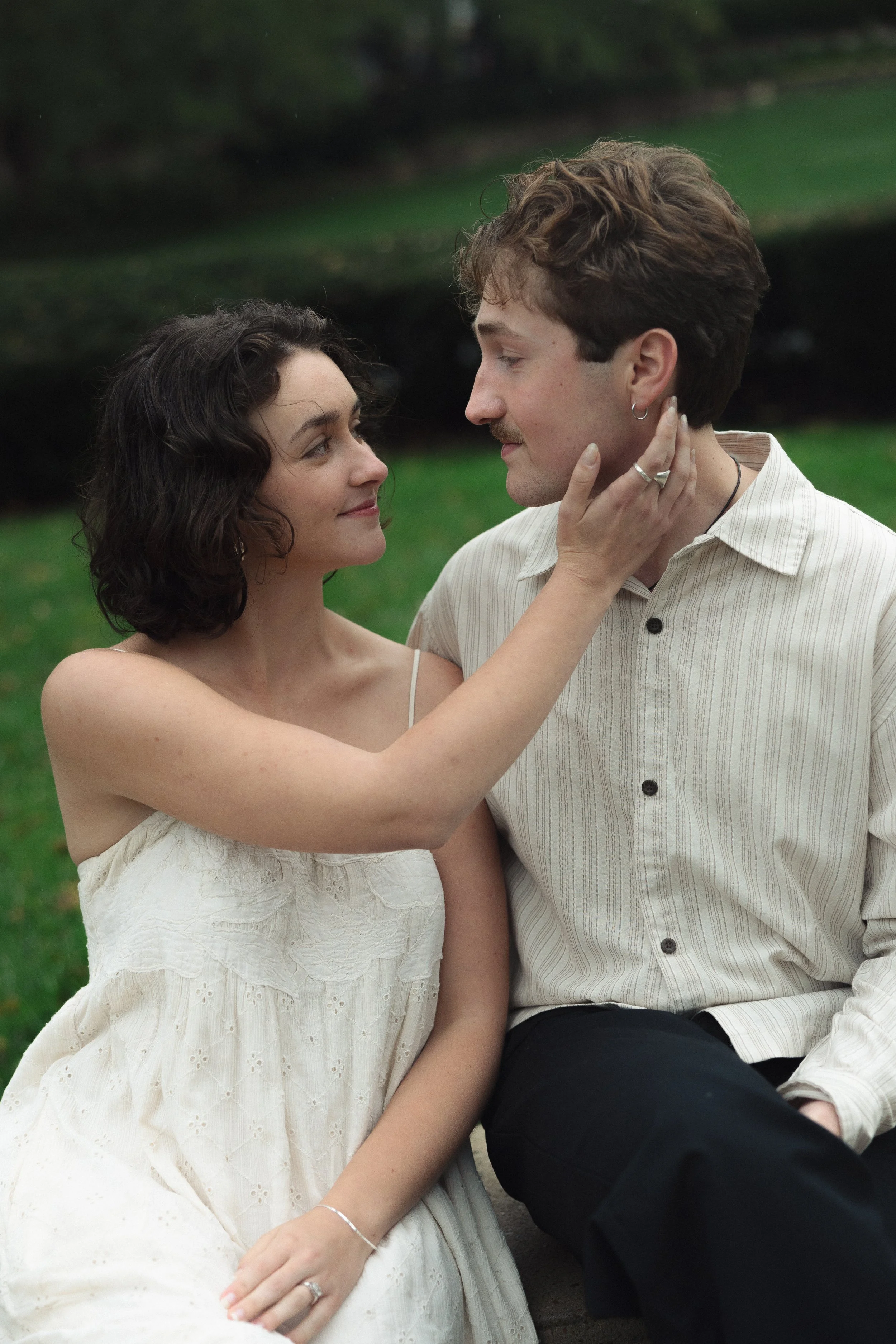 A woman and a man sitting closely outside, gazing at each other. The woman is touching the man's face gently, and they are both smiling softly.