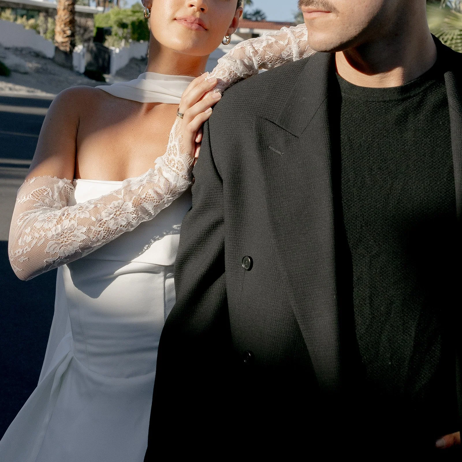 A couple dressed in wedding attire, the woman in a white gown with lace gloves and the man in a black tuxedo, standing outdoors with sunny weather.