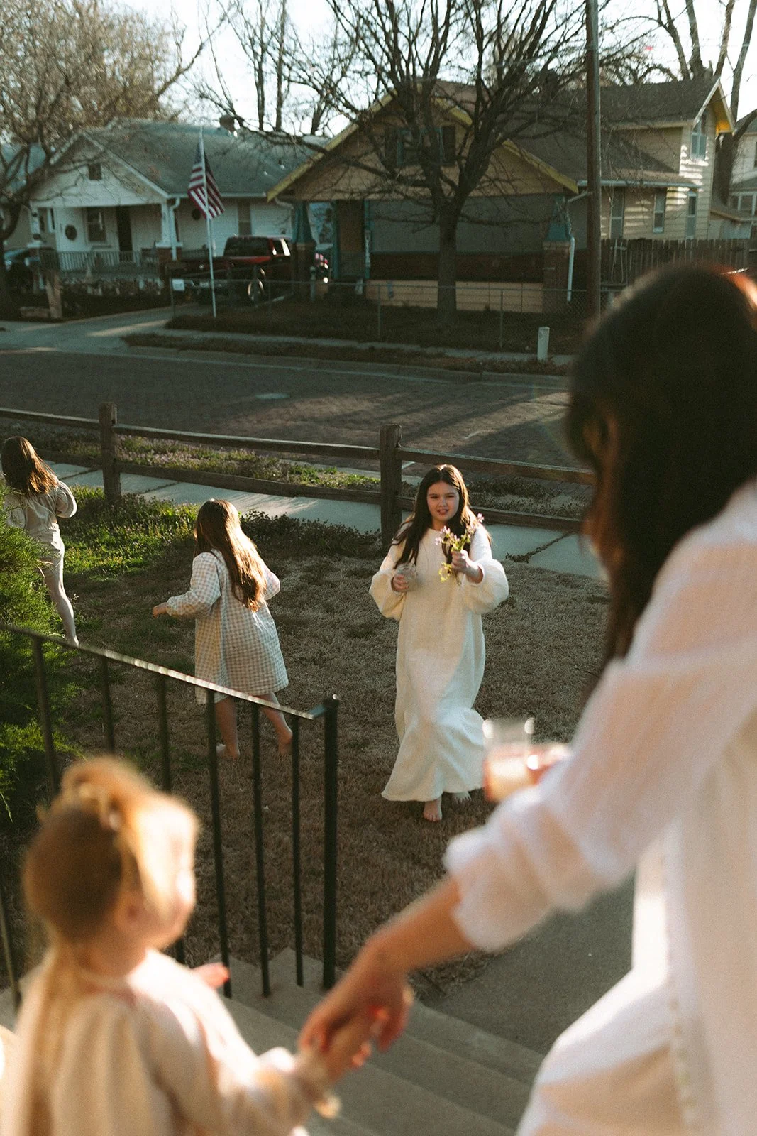 Children playing outside in a yard during the evening, with a girl in a white dress holding flowers and a woman partially visible on a porch, and houses and cars in the background.