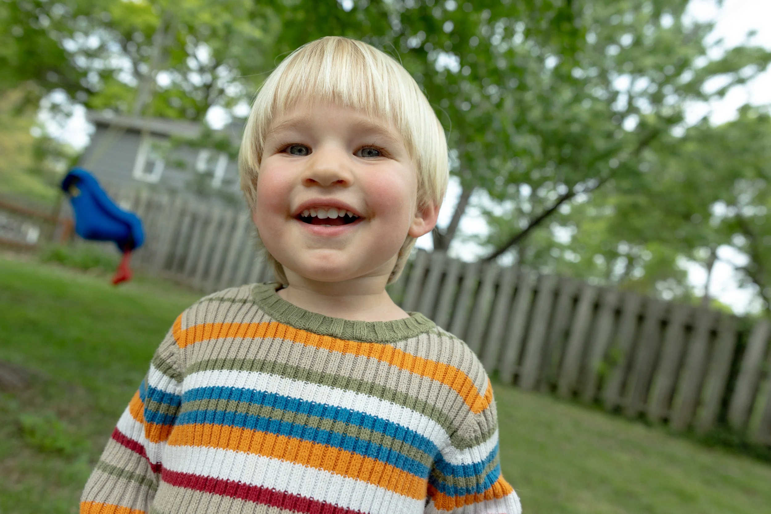 A young boy with blonde hair and a multicolored striped sweater smiling outdoors with a wooden fence and green trees in the background.
