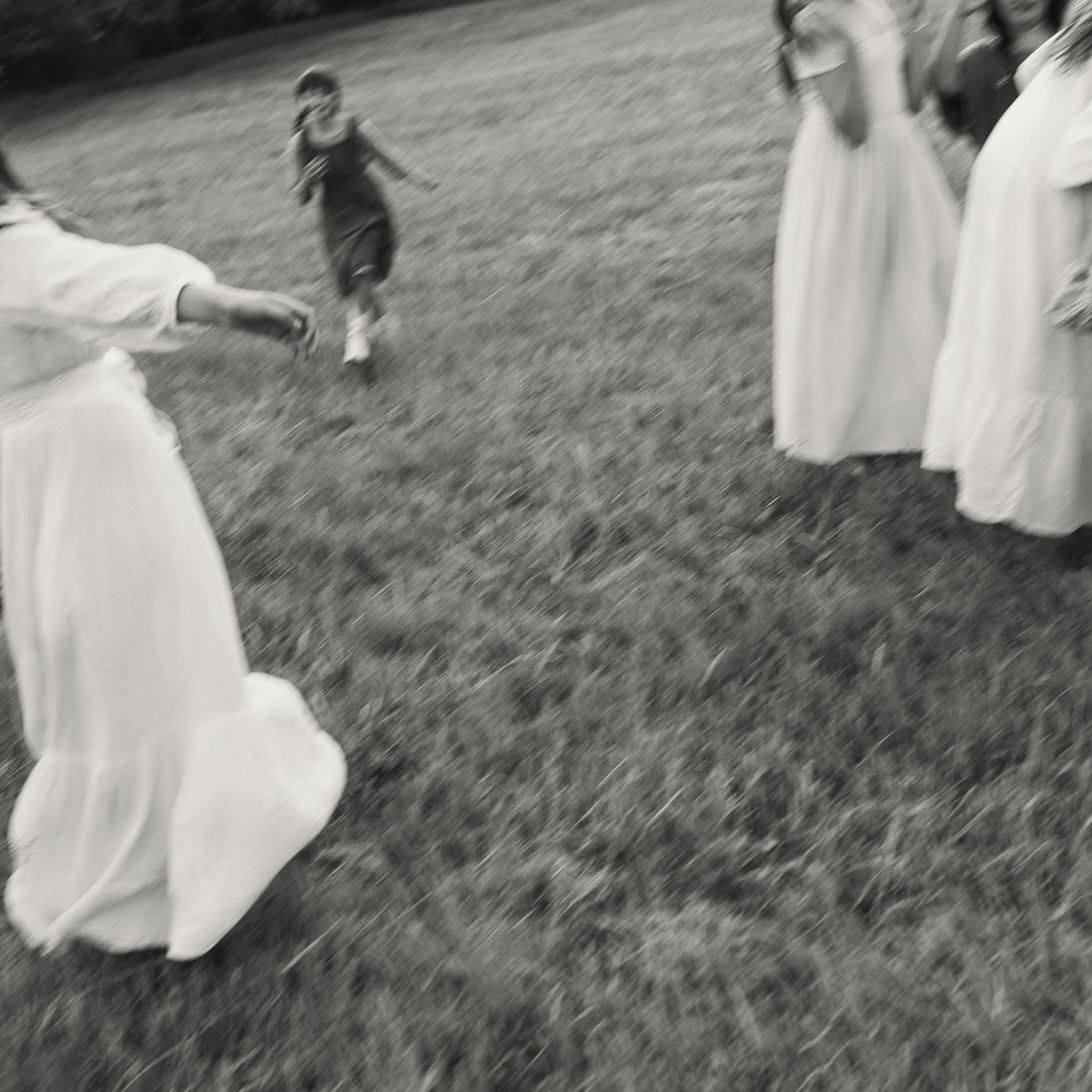 Children playing on a grassy field, one child running toward the camera and others dressed in white dresses standing on the side.