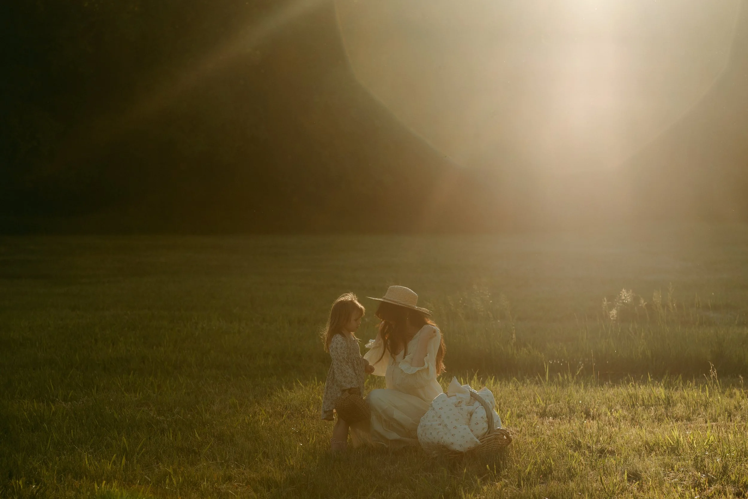 A woman in a white dress and sun hat kneels on the grass, talking to a young girl in a patterned dress, with a basket and blankets nearby, during sunset in a field.