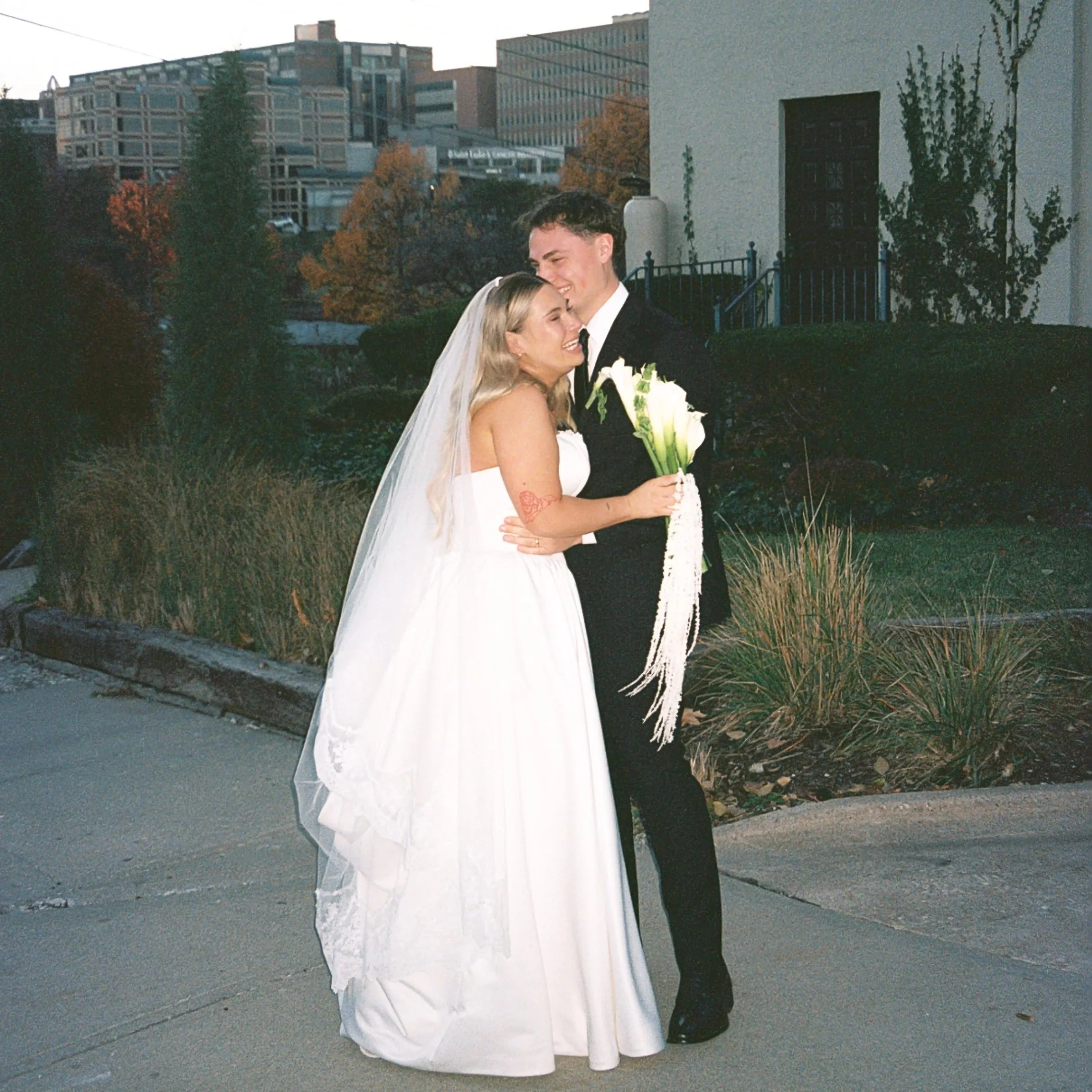 A newlywed couple dressed in wedding attire, smiling and embracing outdoors during sunset, with urban buildings and landscaped garden in the background.