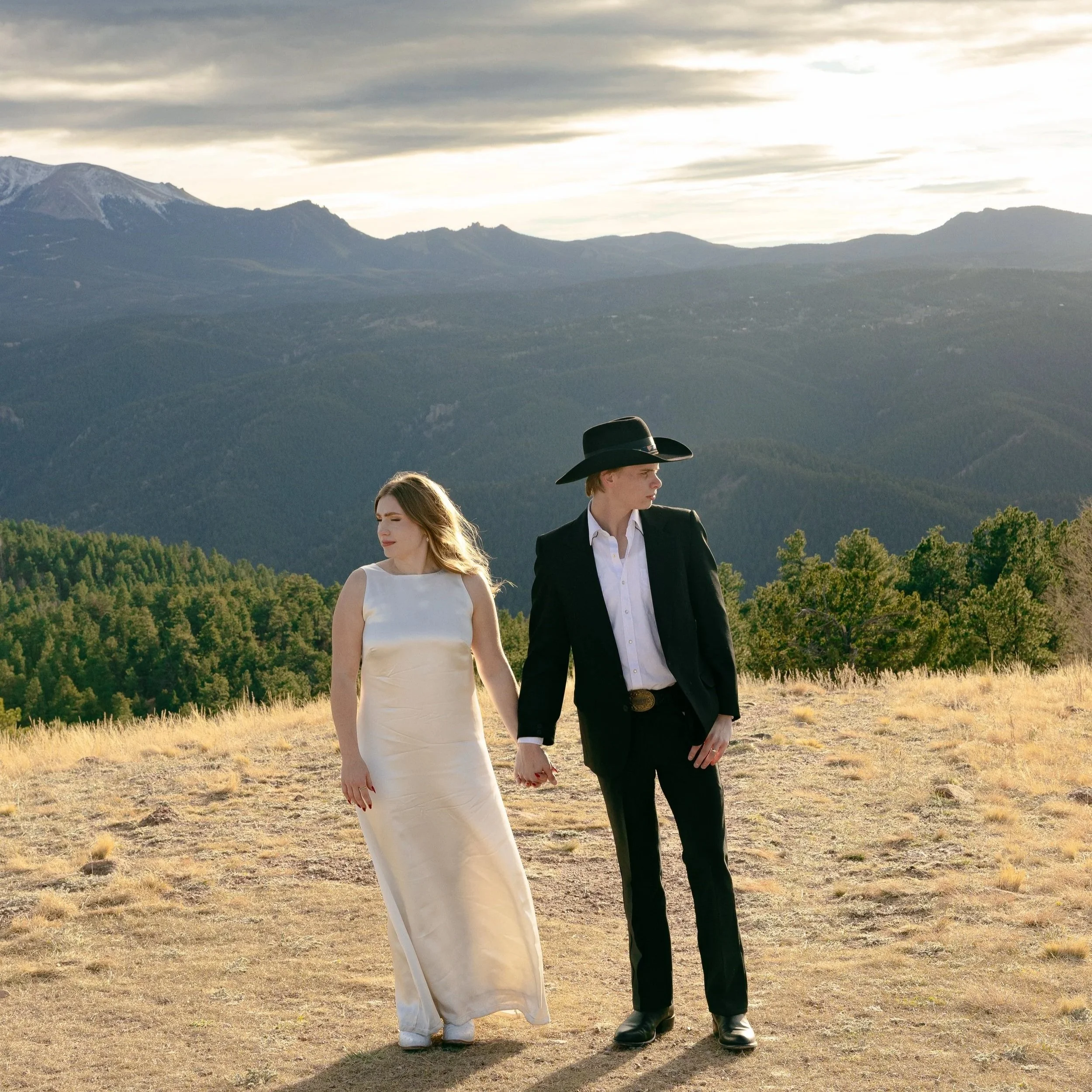 A man and woman holding hands in a field with mountains in the background, the woman wearing a white dress and the man in a black suit and cowboy hat.