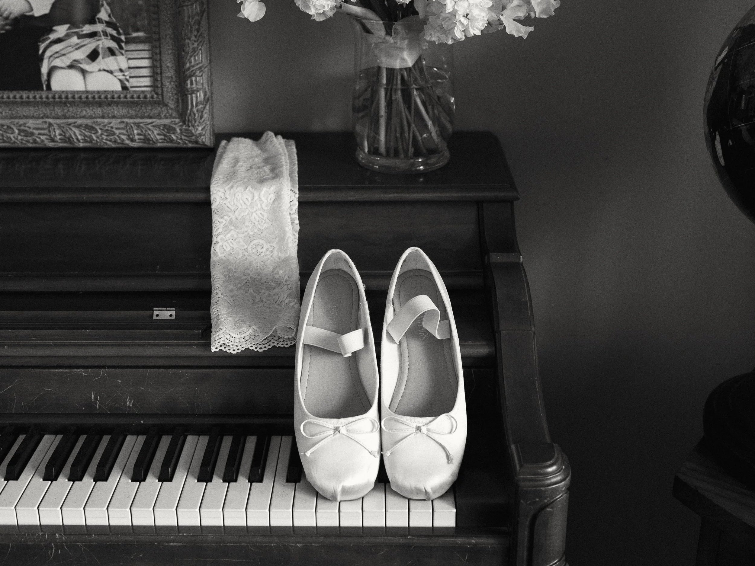White ballet flats with bows on a piano keyboard, candles, lace cloth, and a vase with flowers on a dark wooden table.