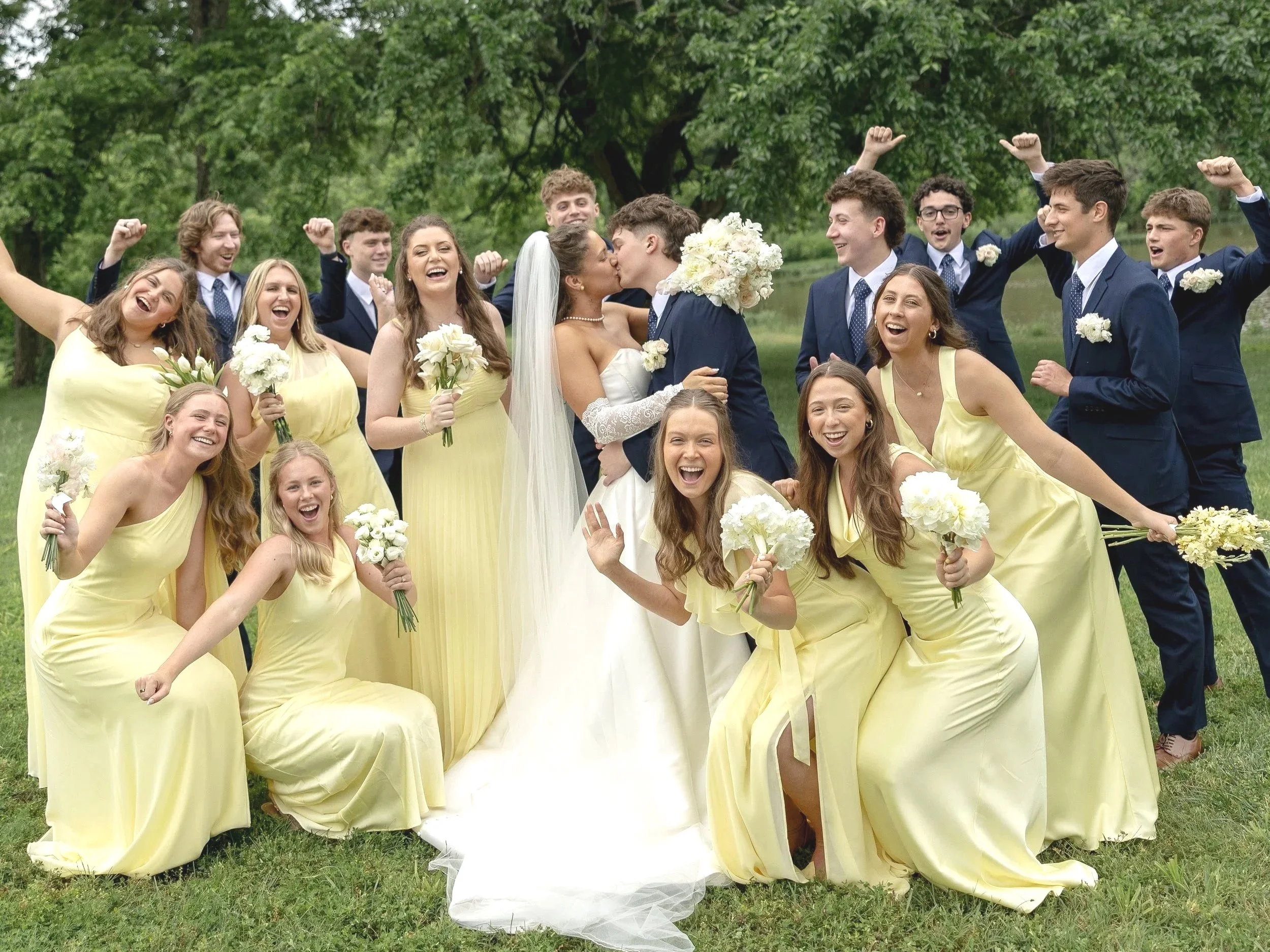 A group of wedding guests, mostly young women in yellow dresses and young men in navy suits, celebrating outdoors near a lake with trees in the background. Two women are kissing in the center, one in a wedding dress and the other in a navy suit, surr