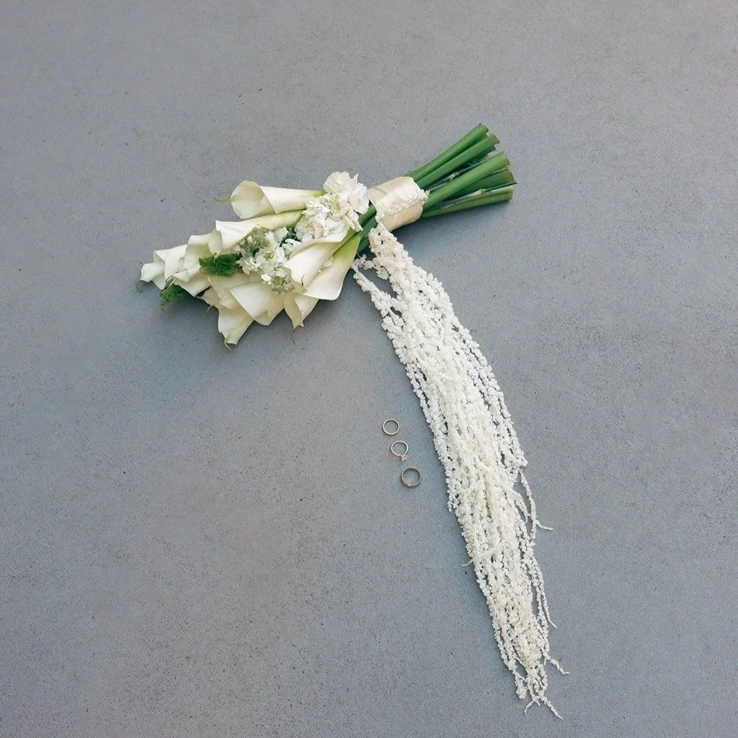A white floral bouquet with green stems, three rings, and a trail of white beads or small flowers on a gray surface