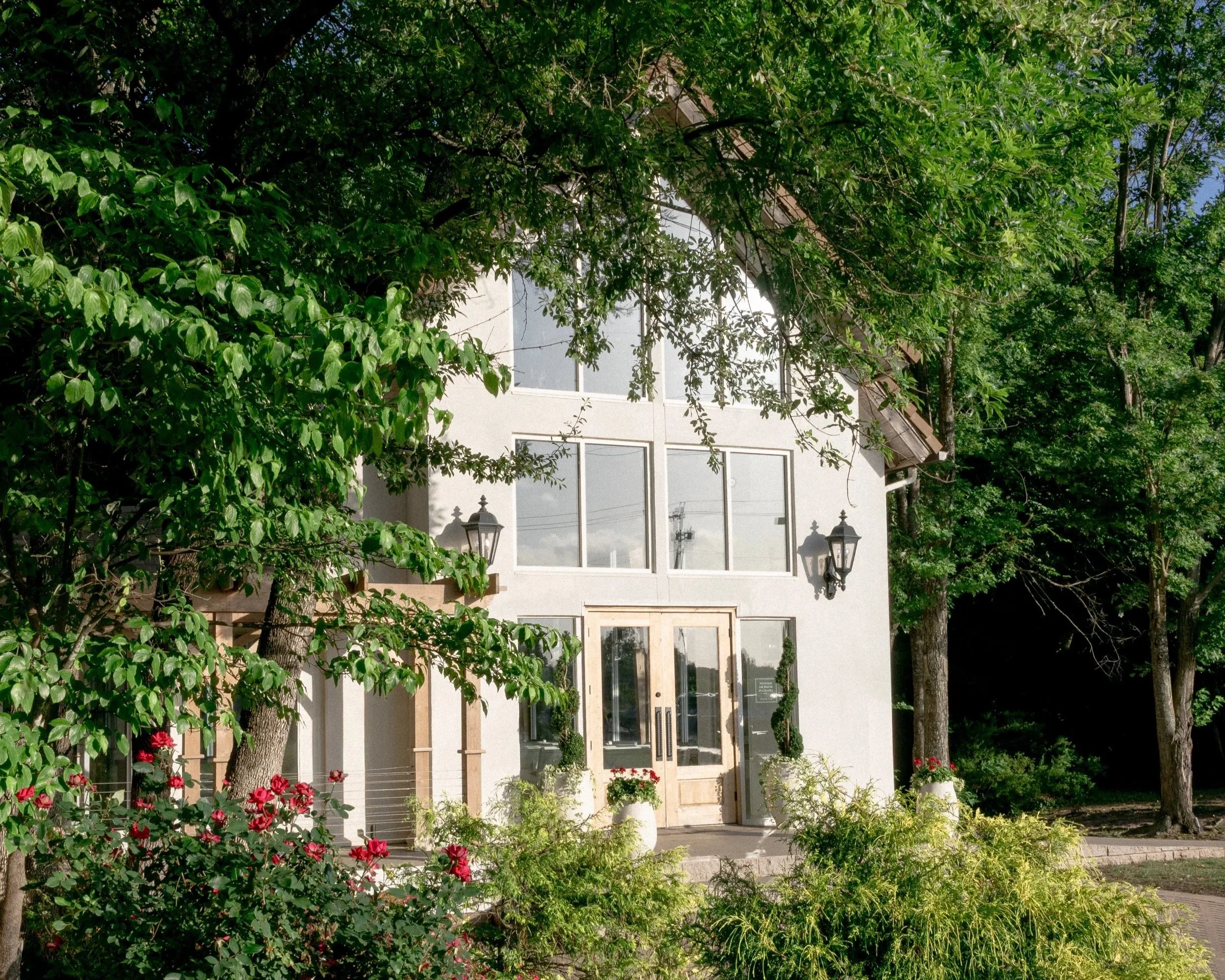 View of a house with large windows and a glass door, surrounded by green trees, plants, and flowers outside.
