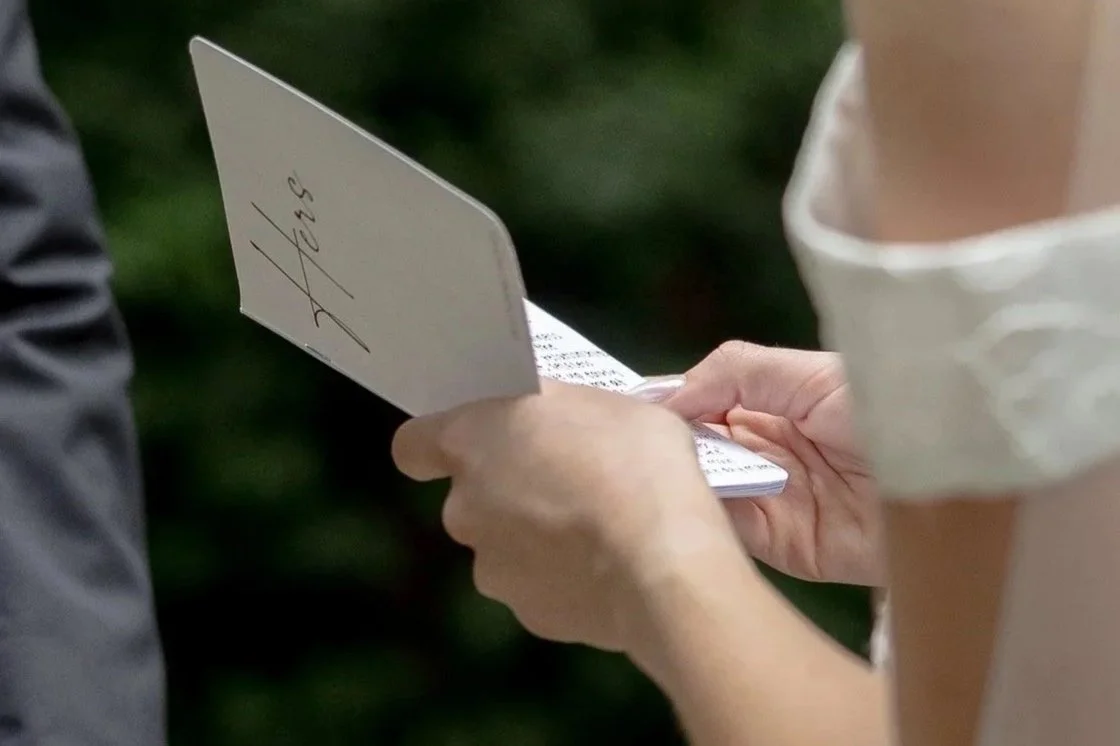 Close-up of a person in a white shirt holding a small notepad with handwritten wedding vows, signing or writing, while another person stands nearby.