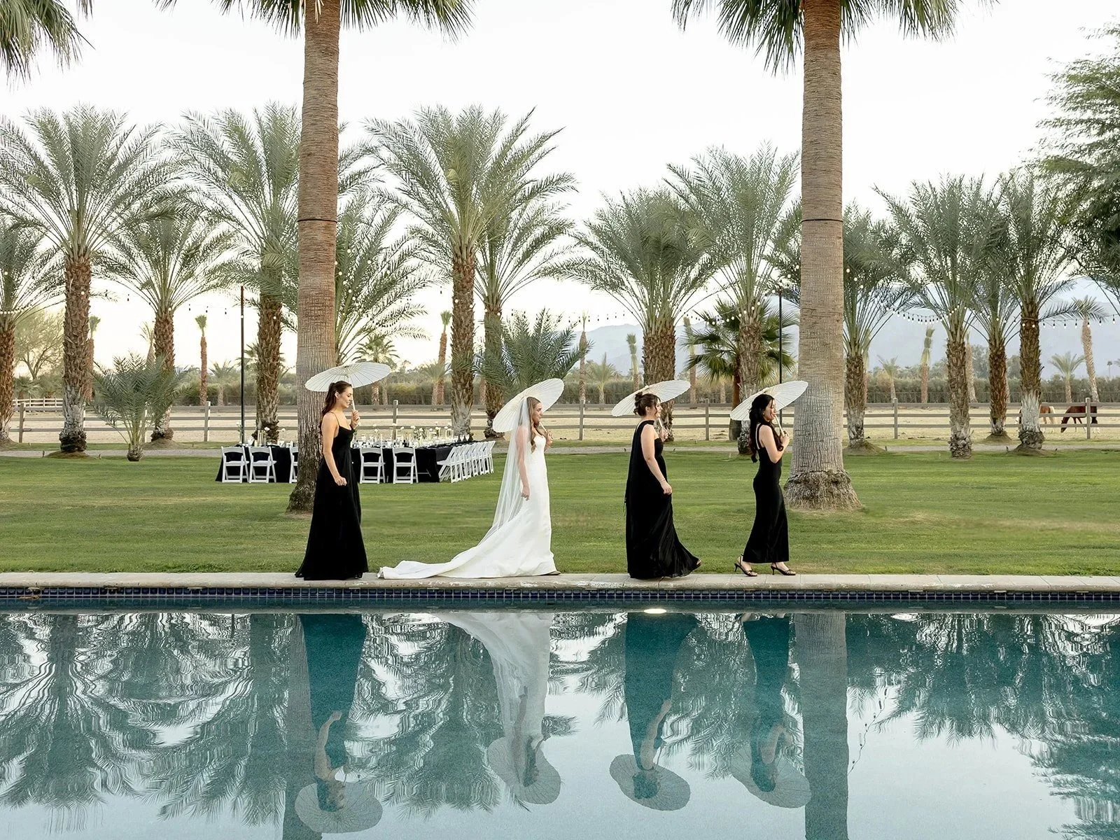 A bride in a white wedding dress and veil, walking on a poolside with four bridesmaids in black dresses holding umbrellas, against a backdrop of palm trees and arranged outdoor dining tables. Palm Spring, California wedding.