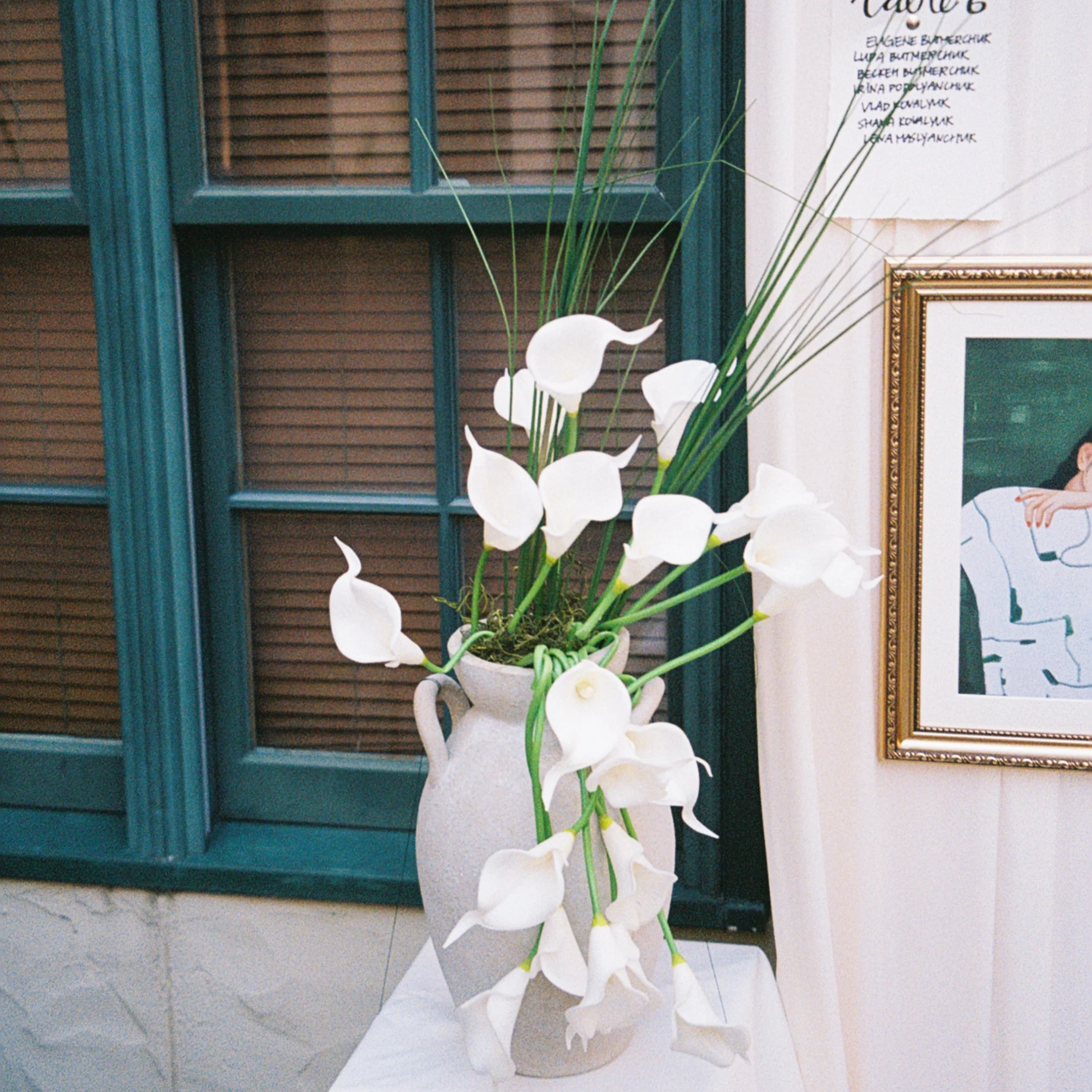 A white ceramic vase with a handle holding white calla lilies and tall green grass against a background of a framed piece of art, a wooden window blind, and a white wall with handwritten notes.