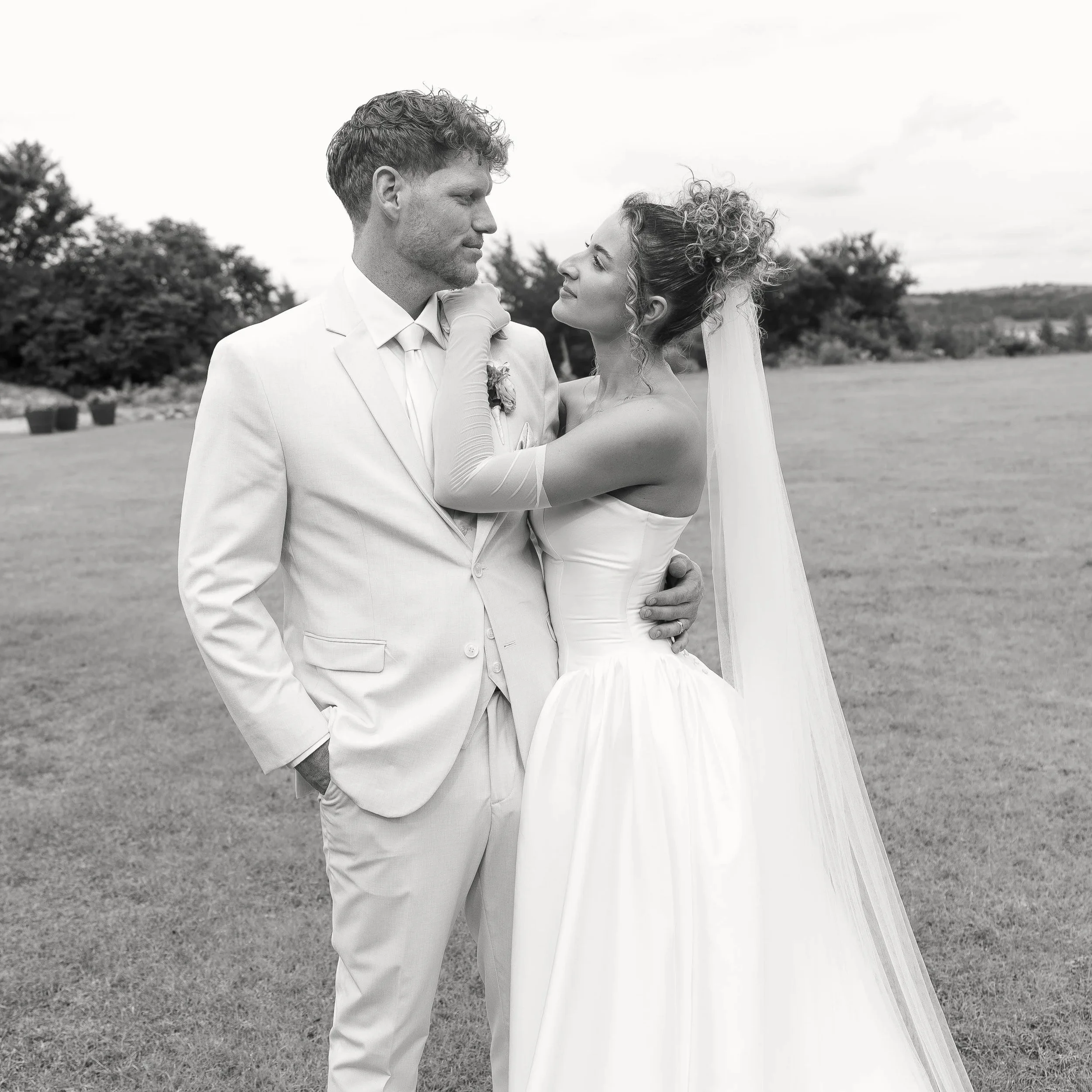 A black-and-white photograph of a couple at their wedding outdoors, with the groom in a white suit and the bride in a strapless wedding gown. They are gazing into each other's eyes, with the bride's arms around the groom's neck, and the groom's hand 
