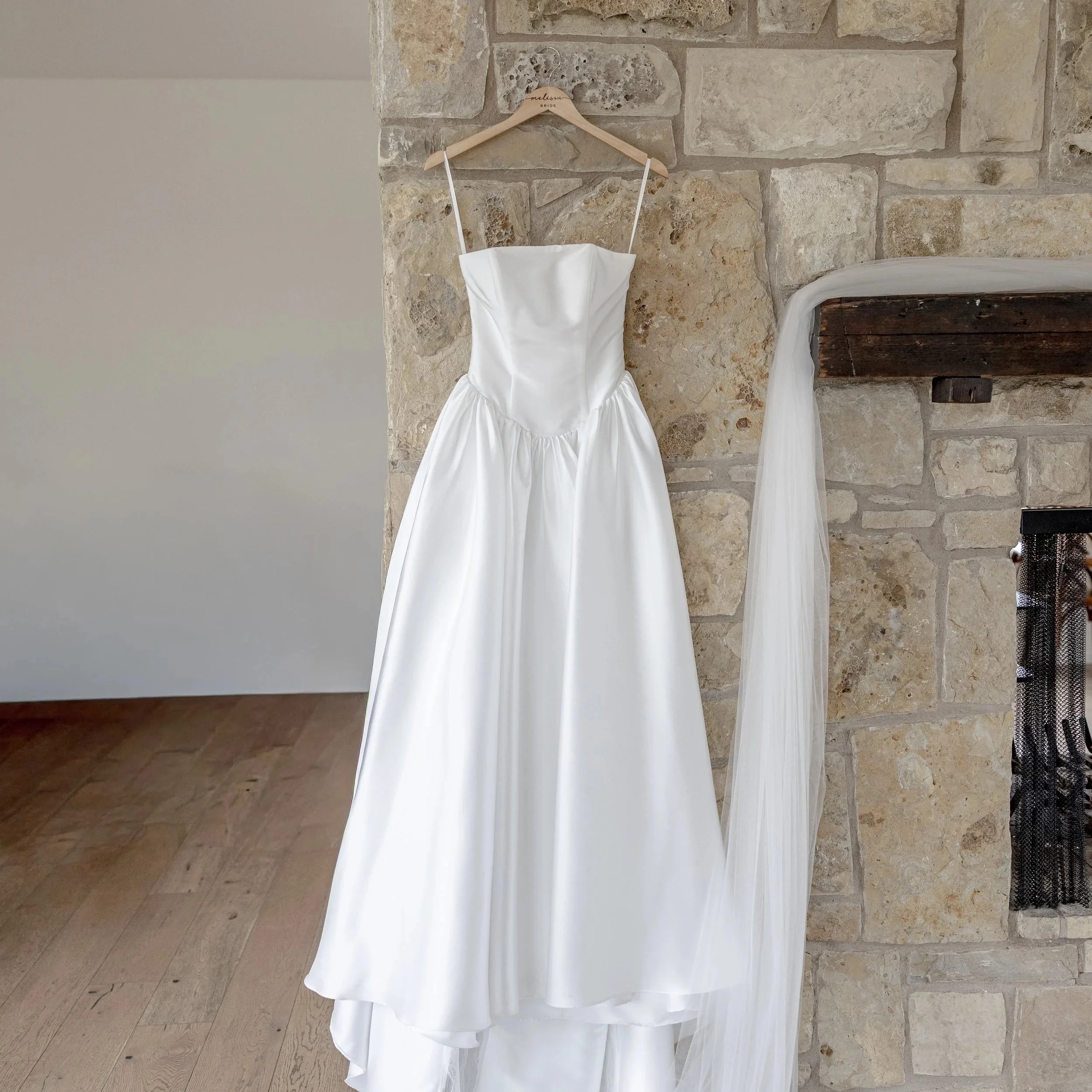 A white wedding dress hanging on a wooden hanger attached to a stone fireplace wall.