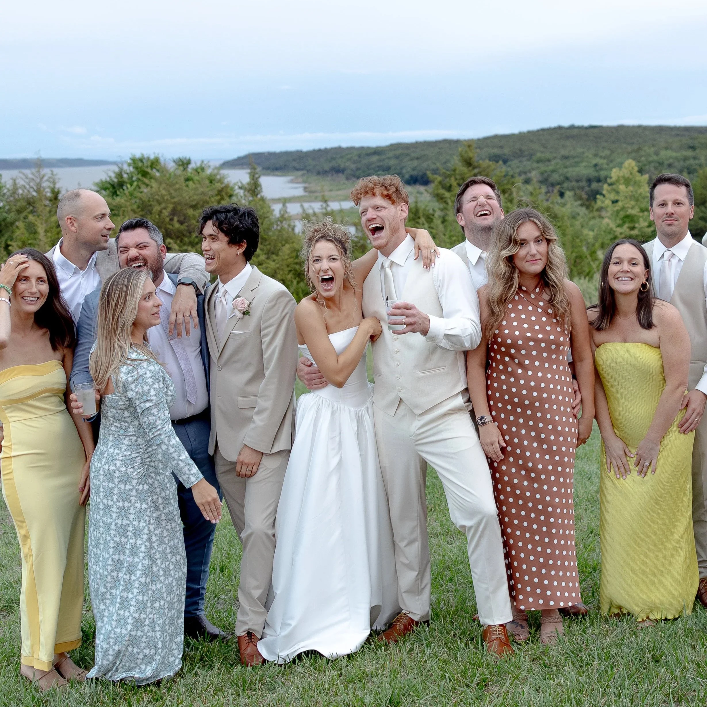 Group of people dressed in formal attire at an outdoor event near a body of water with trees in the background. They are smiling and laughing, with some showing excitement or joy.