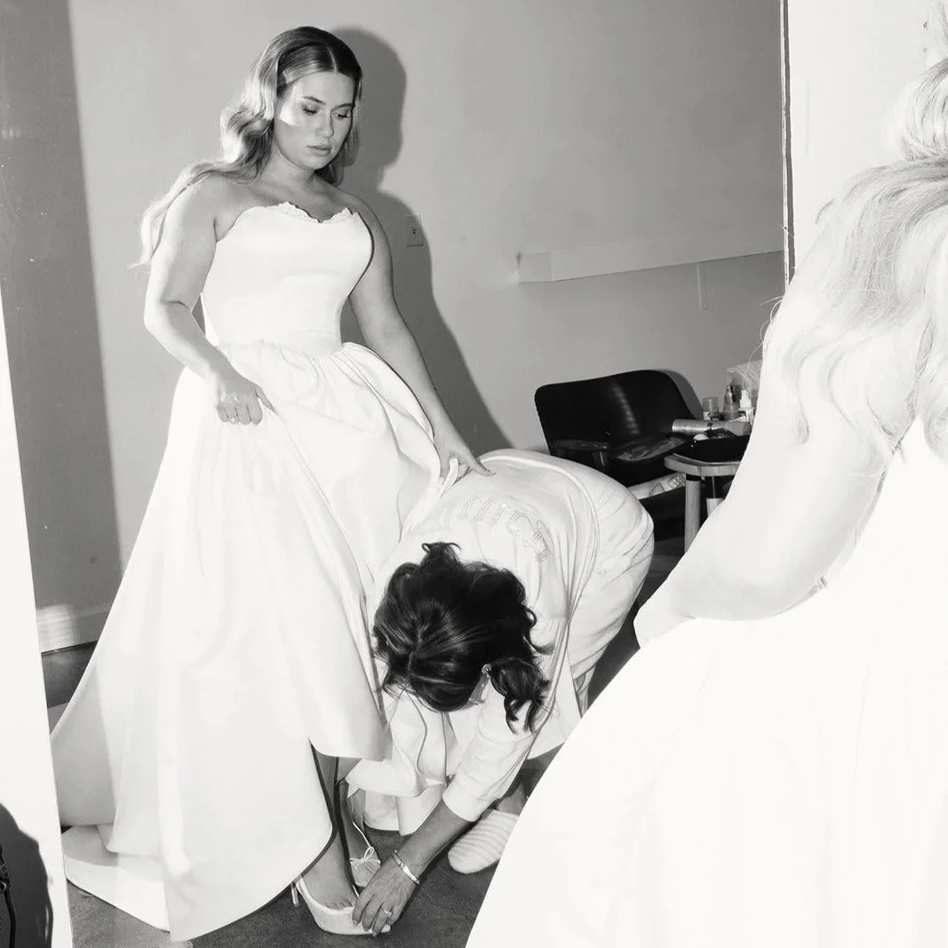 A woman in a wedding dress looks down at another person adjusting her shoe while another woman stands nearby in a room.
