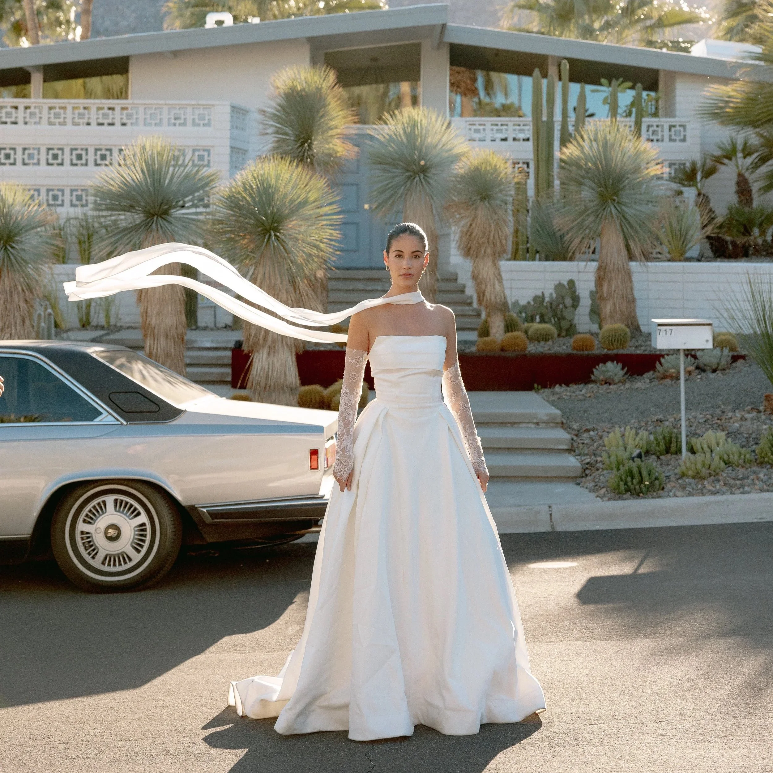 A woman in a white wedding dress stands outdoors in front of a modern house with a white exterior, surrounded by desert plants including cacti and yuccas. The scene is illuminated by sunlight, and a vintage car is parked nearby.