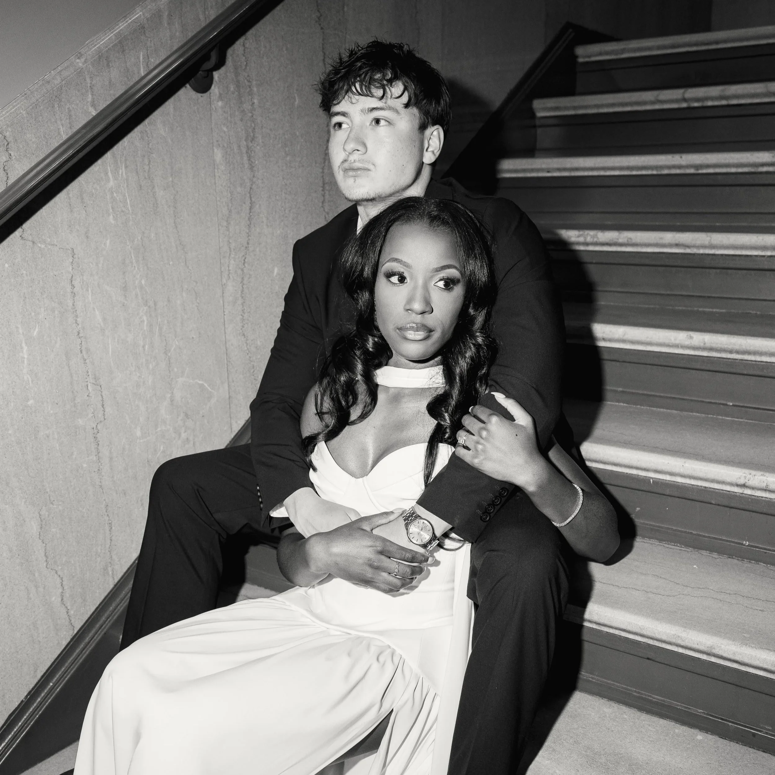 A black-and-white photo of a young man and woman sitting together on a staircase. The man has wavy hair, light skin, and is looking away with a serious expression, wearing a dark suit. The woman has wavy hair, darker skin, and is looking slightly to 