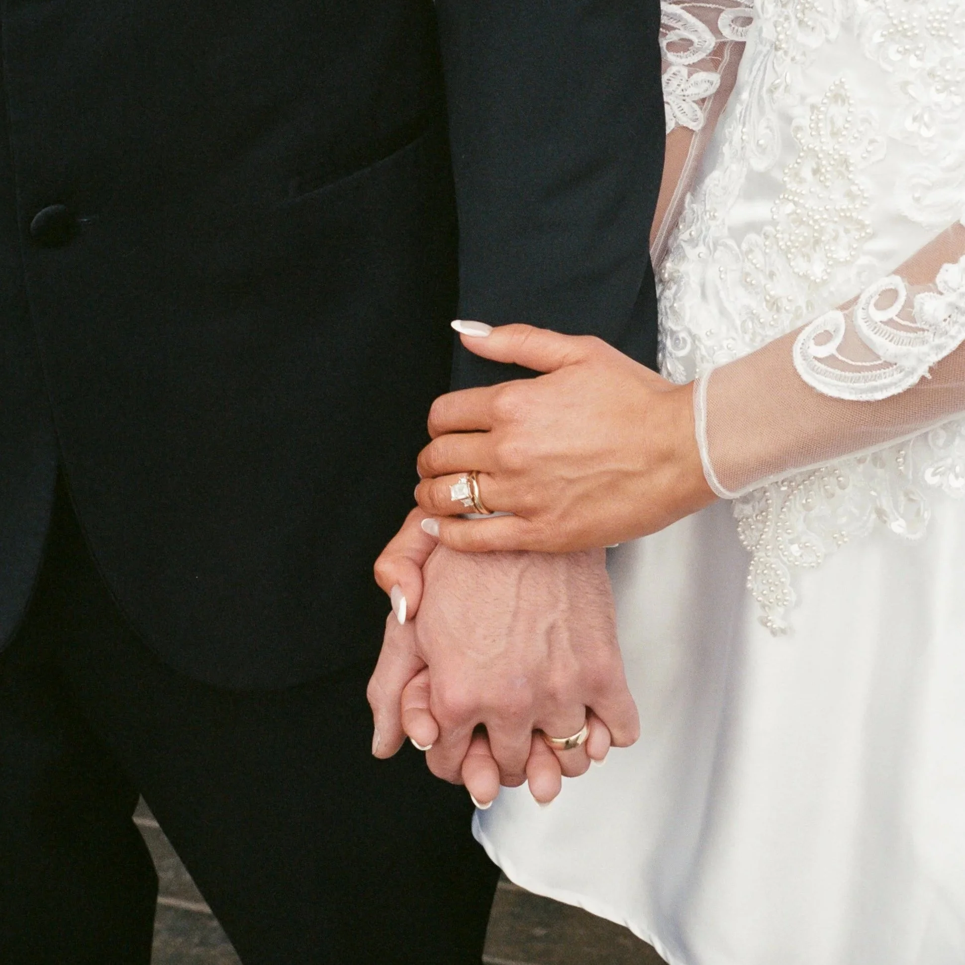 Close-up of two people holding hands, one wearing a black suit and the other in a white lace wedding dress, with their hands intertwined.