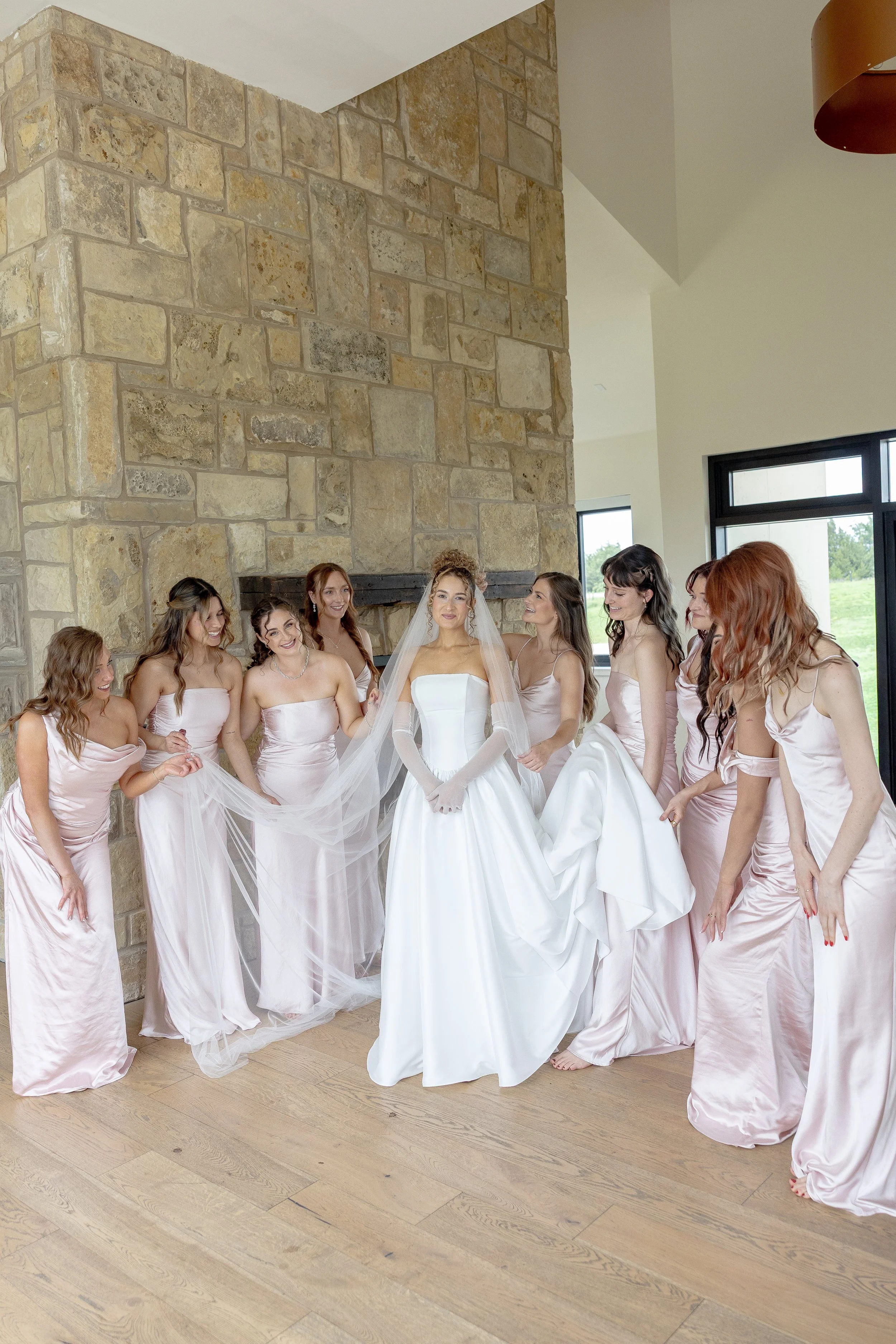 A bride in a white wedding dress is surrounded by bridesmaids in matching pink satin dresses inside a room with a stone fireplace and large windows.