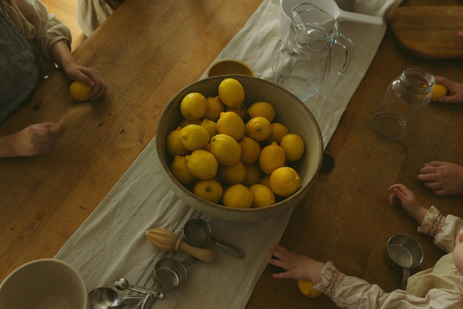 A top-down view of a wooden dining table with a white cloth runner, a large bowl filled with yellow lemons, glass pitchers, measuring spoons, and children's hands reaching for the lemons.