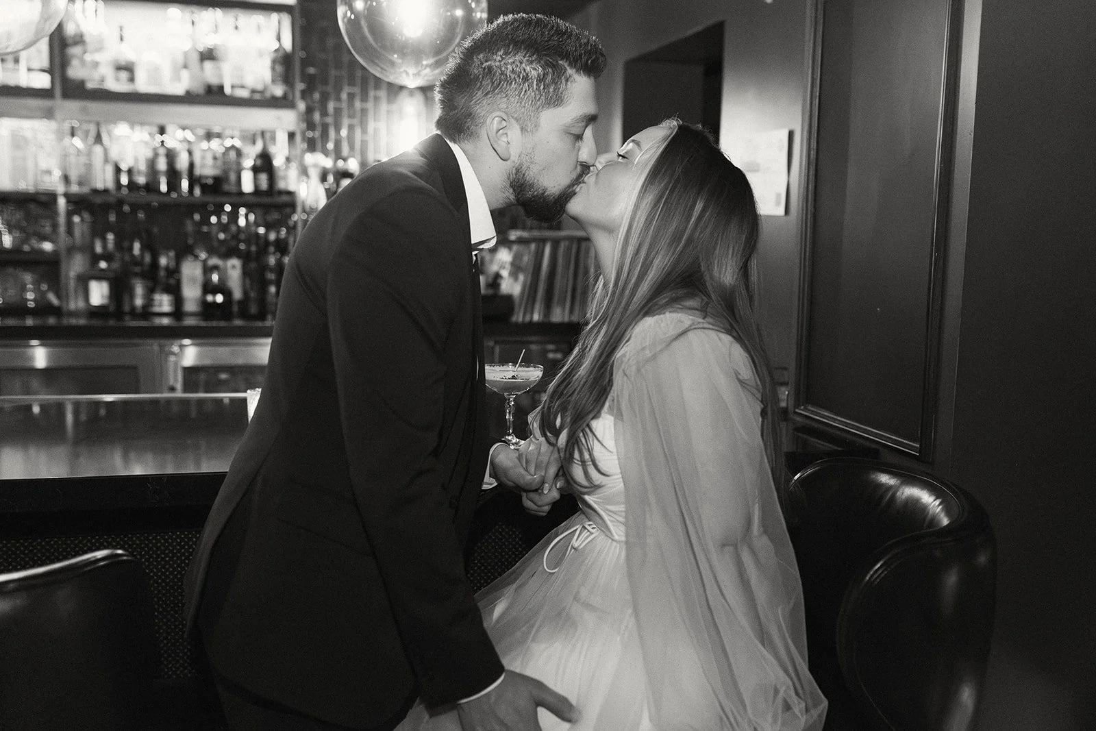 A man in a tuxedo and a woman in a dress sharing a kiss in a bar.